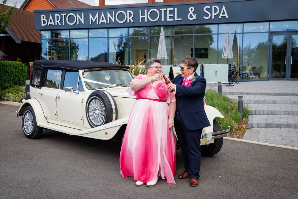 Couple embracing in pink and navy attire outside Barton Manor Hotel & Spa beside a vintage cream wedding car.
