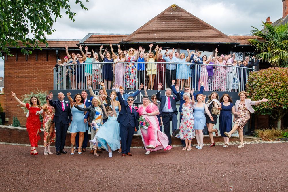 Large wedding party celebrating outdoors with guests cheering on a balcony behind the happy couple.