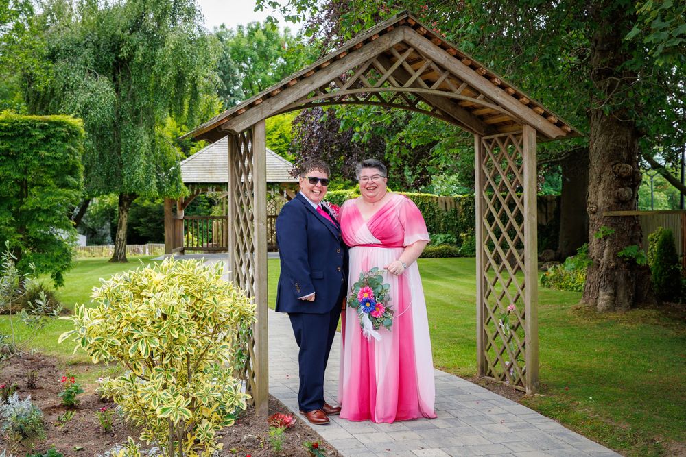 Couple posing under wooden garden arbor, woman in pink gown, man in navy suit, lush green garden backdrop.