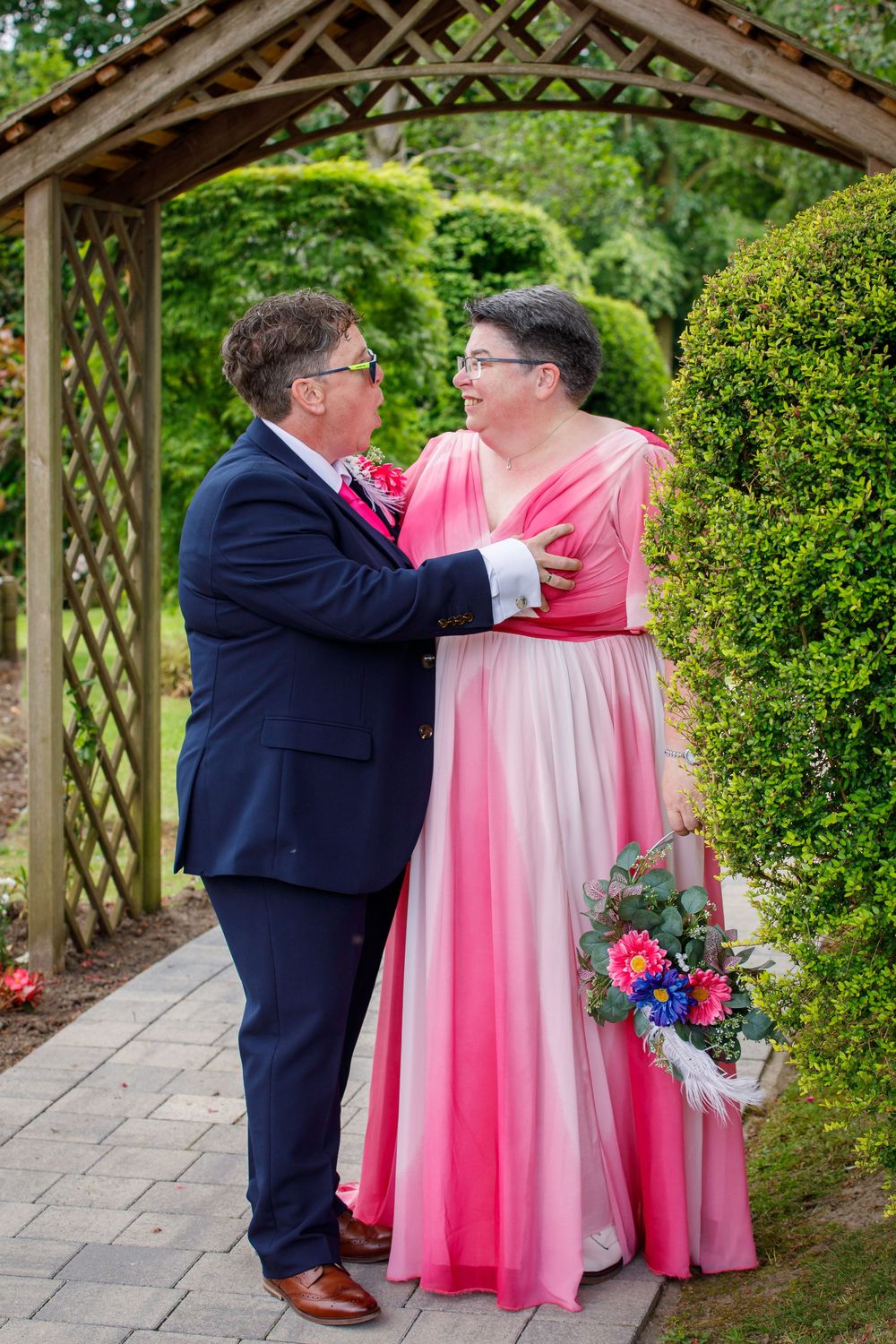 Couple in formal attire, navy suit and pink ombre gown, share a tender moment under a wooden garden arbor.
