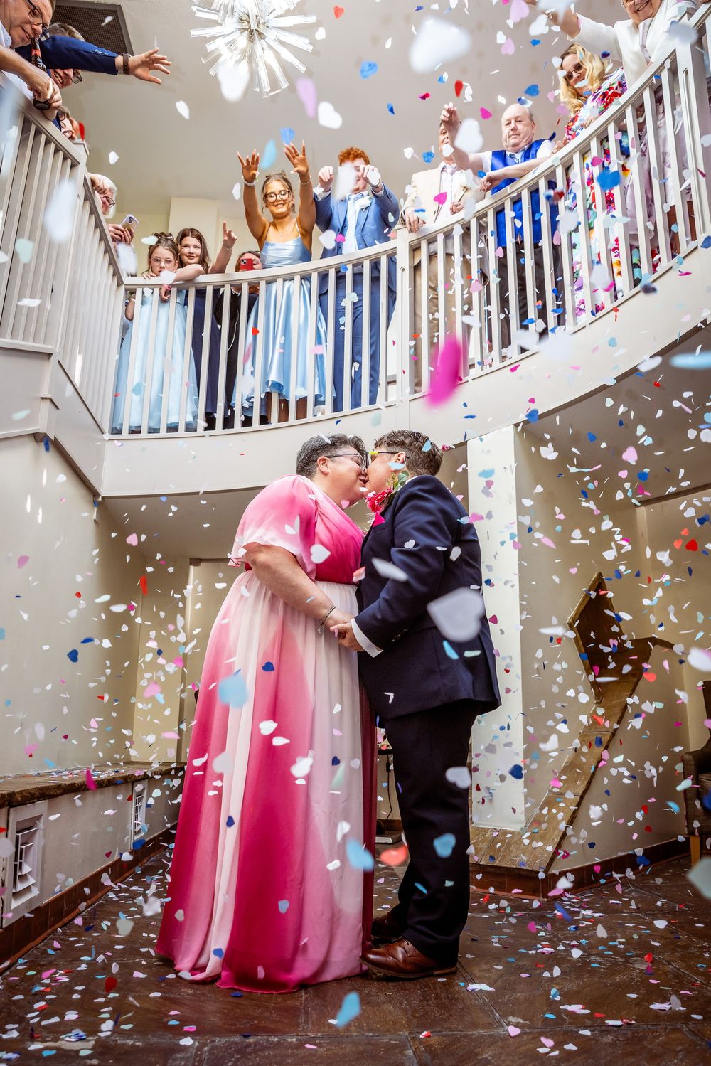 Two brides share a kiss under colorful confetti as guests celebrate on a staircase balcony above them.