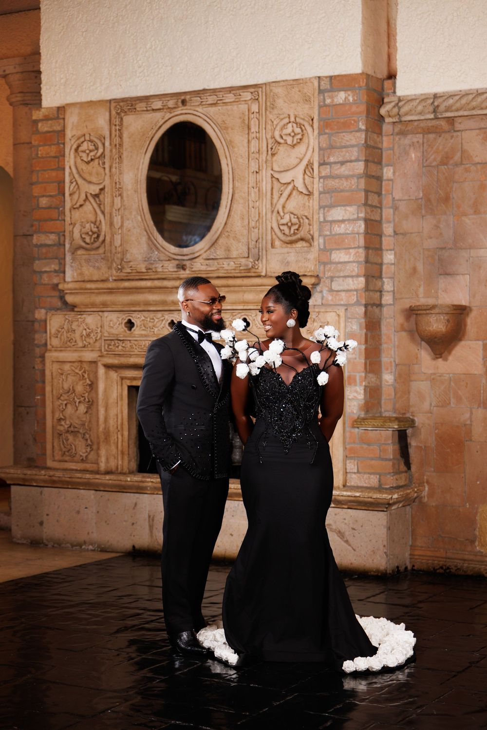Elegant Black couple in formal attire pose before ornate brick fireplace, woman in black gown with white flowers.