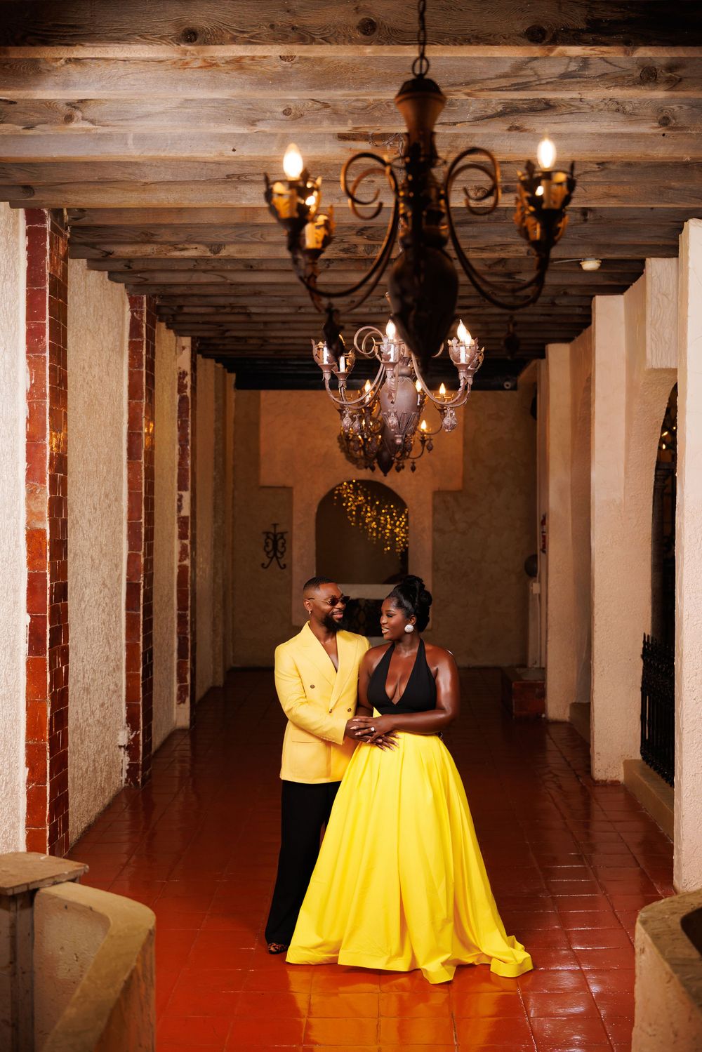 Elegant couple in yellow gown and gold blazer posing under chandelier in rustic venue with brick walls.