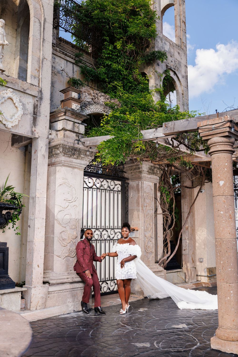 Elegant couple in wedding attire posing before ornate stone columns with lush greenery at a historic venue.