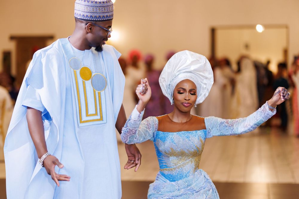 Couple dancing in traditional Nigerian attire, man in blue agbada, woman in lace dress with gele headwrap.