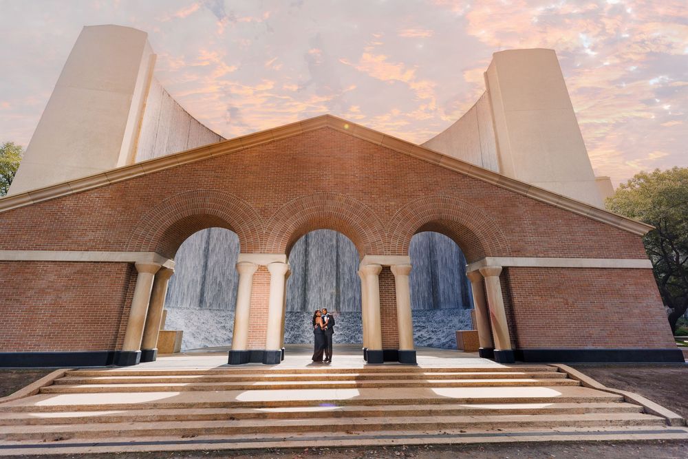 Couple poses before a grand brick building with arched entrances and tall columns at sunset.