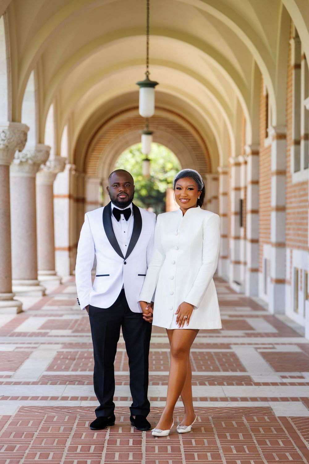 Elegant couple in matching white outfits posing in a beautiful arched corridor with brick flooring.