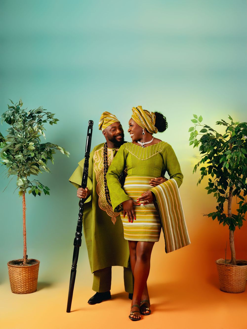 Couple in matching olive green African traditional attire posing together with a walking stick and potted plants.