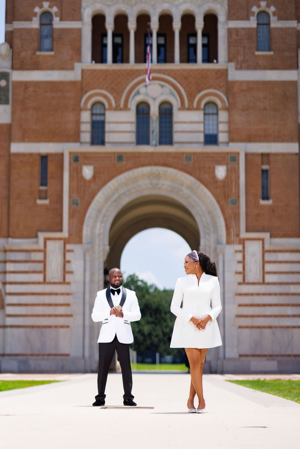 Couple in white formal attire posing in front of a grand brick archway building on a sunny day.