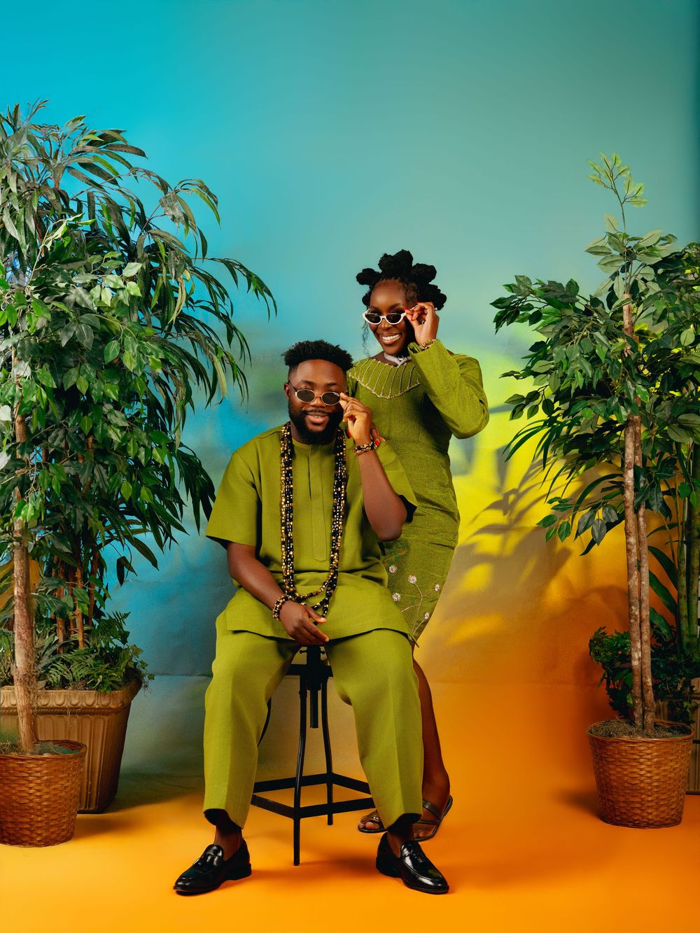 Two people in matching green outfits pose with plants against a teal and yellow background.