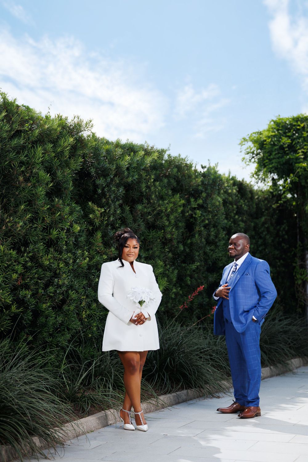 A bride in a white blazer dress holds a rose while a groom in a blue suit stands beside lush green hedges.