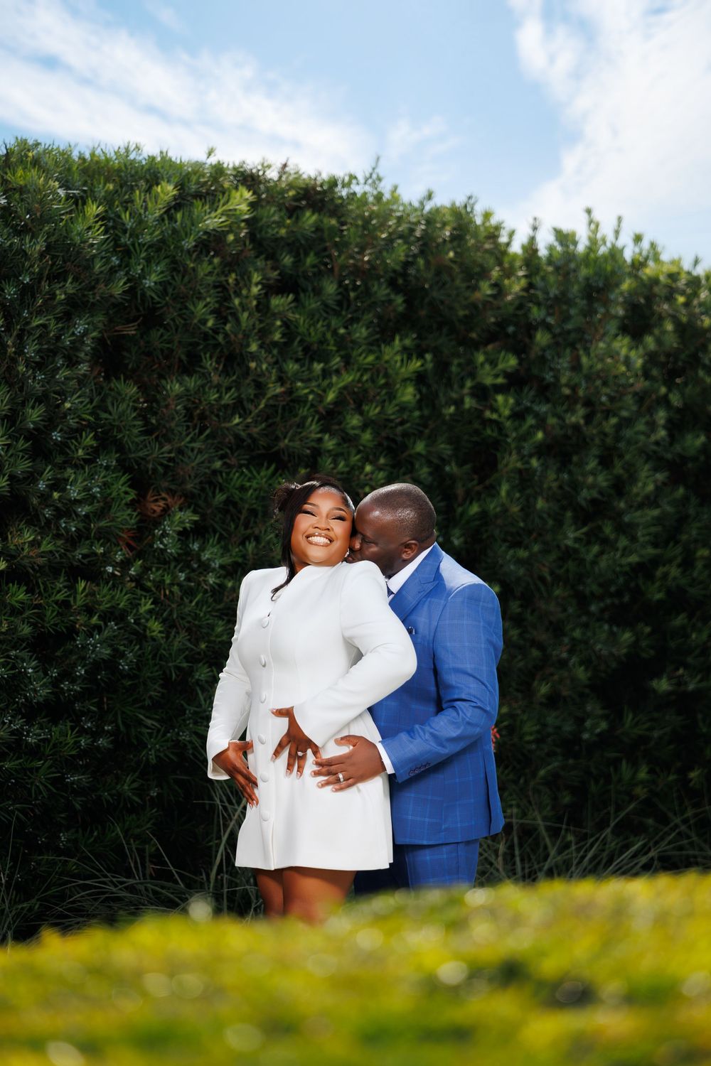 A couple poses in a garden, woman in white dress smiling as man in blue suit embraces her from behind.