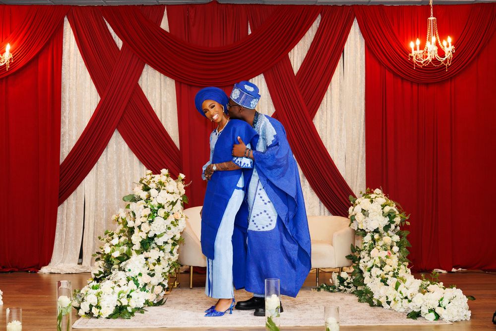 Two people in royal blue traditional African attire embrace before a red and white draped backdrop with floral arrangements.