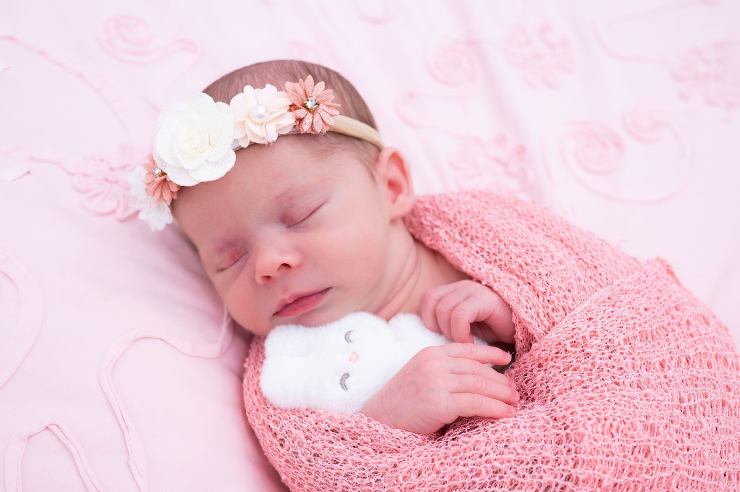 Newborn baby girl sleeping swaddled in pink wrap with floral headband on pink background.