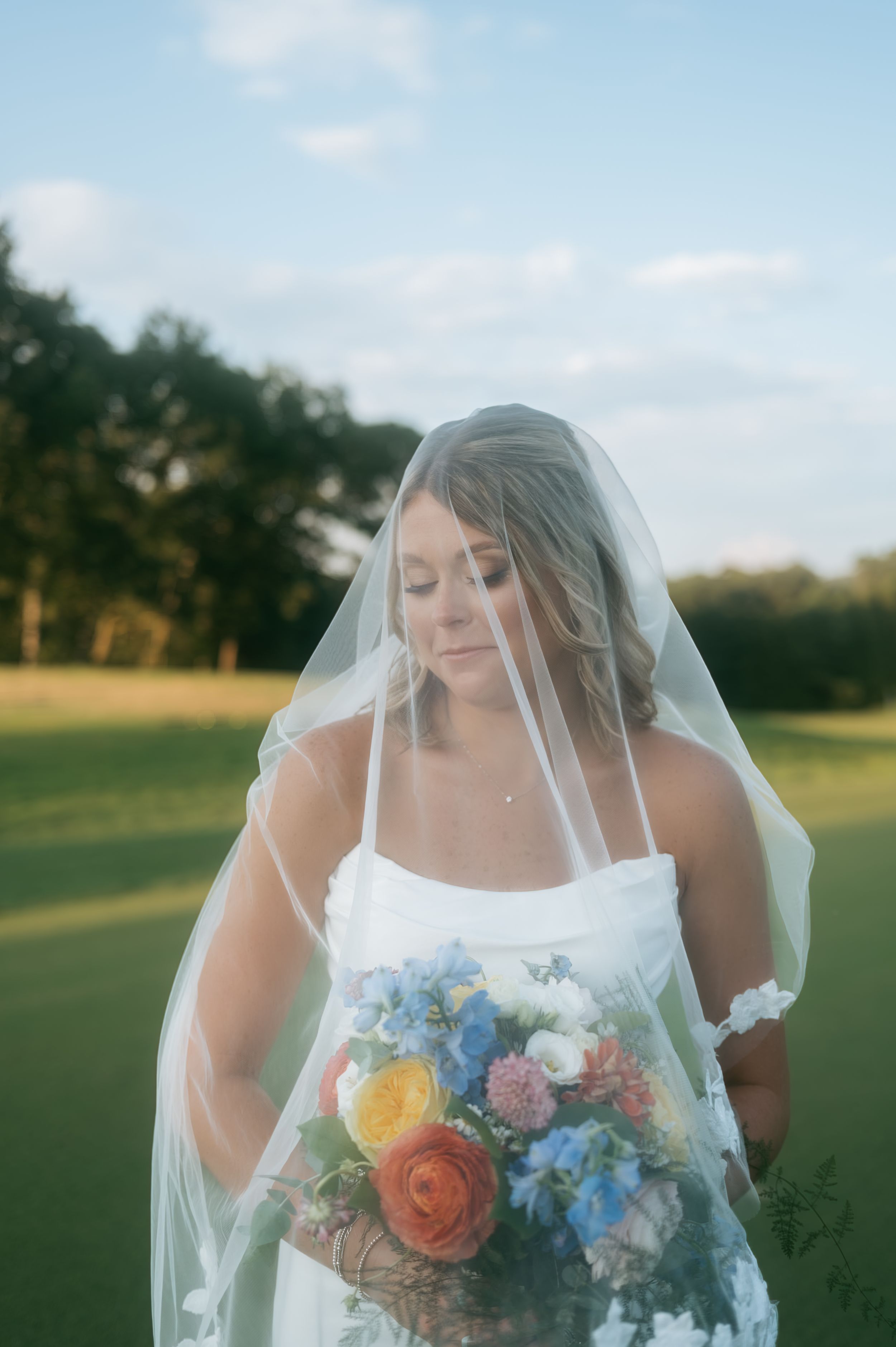 Elegant bride in white gown and veil gazes down at colorful bouquet on a sunlit golf course, wedding photographer Greensboro NC