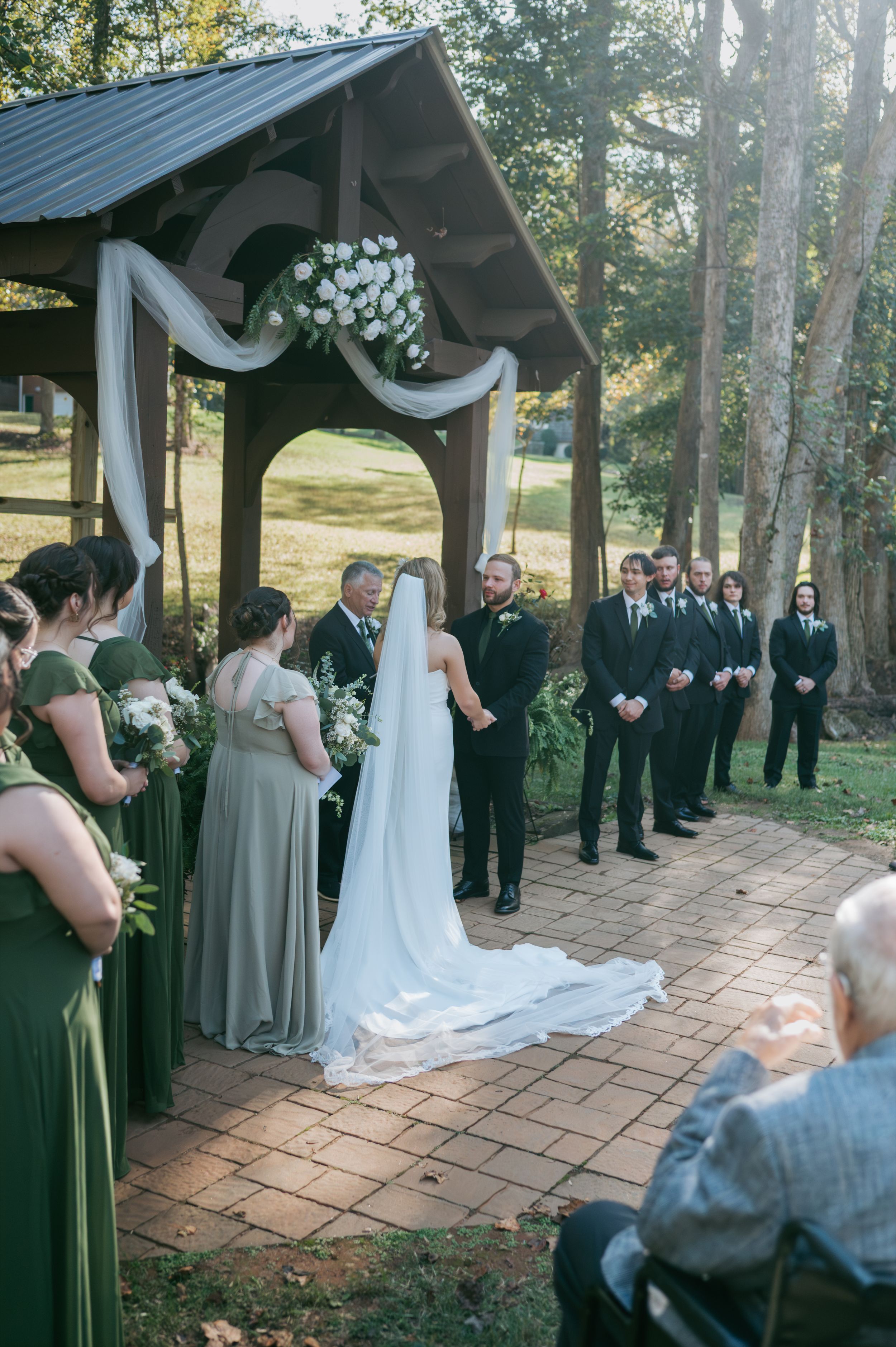 Wedding ceremony at rustic outdoor gazebo with bride, groom, groomsmen in black, and bridesmaids in sage green, Wedding photographer NC