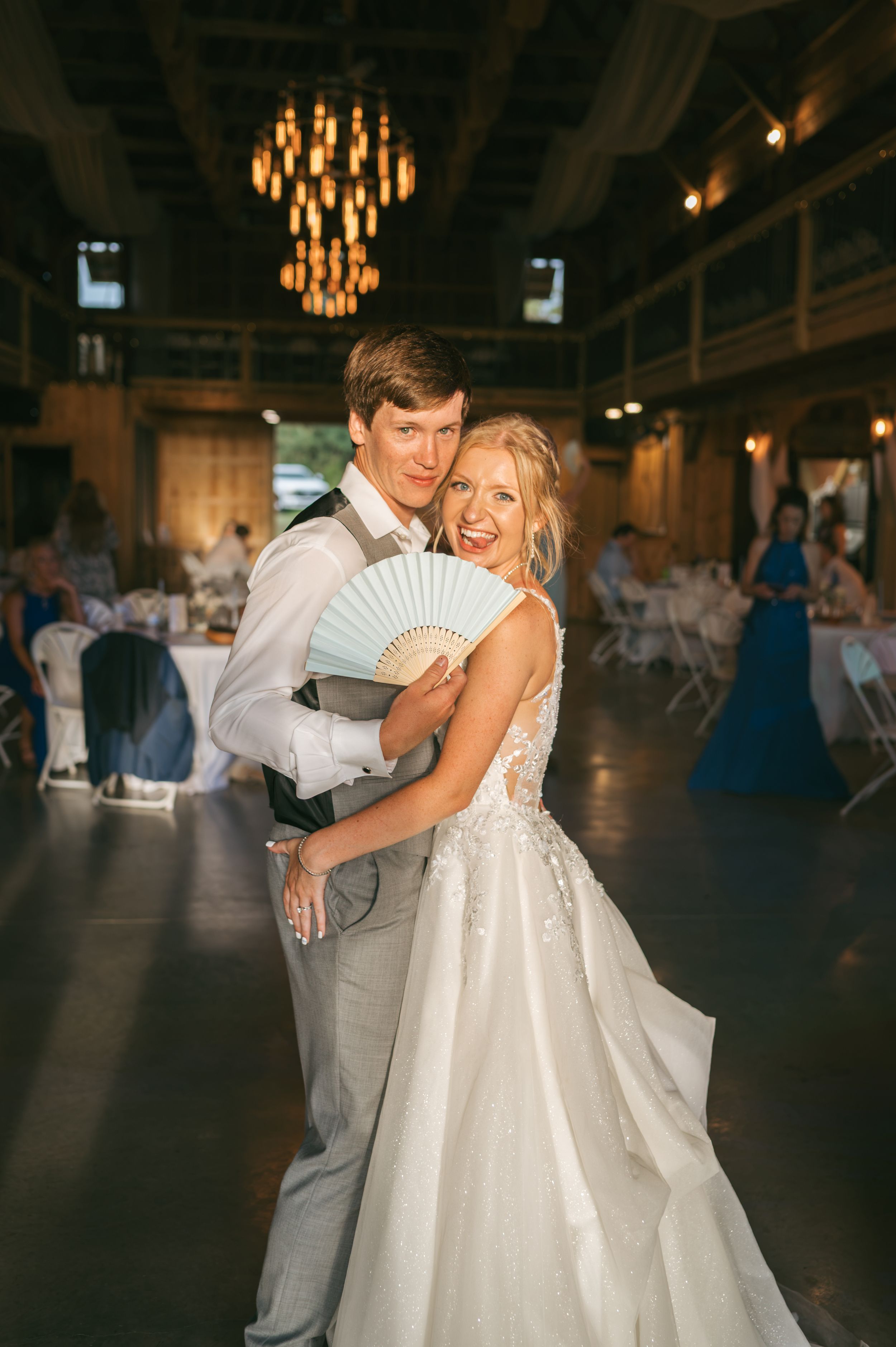 Smiling bride holding a blue fan embraces groom on dance floor at elegant indoor wedding reception with chandelier, wedding photographer NC