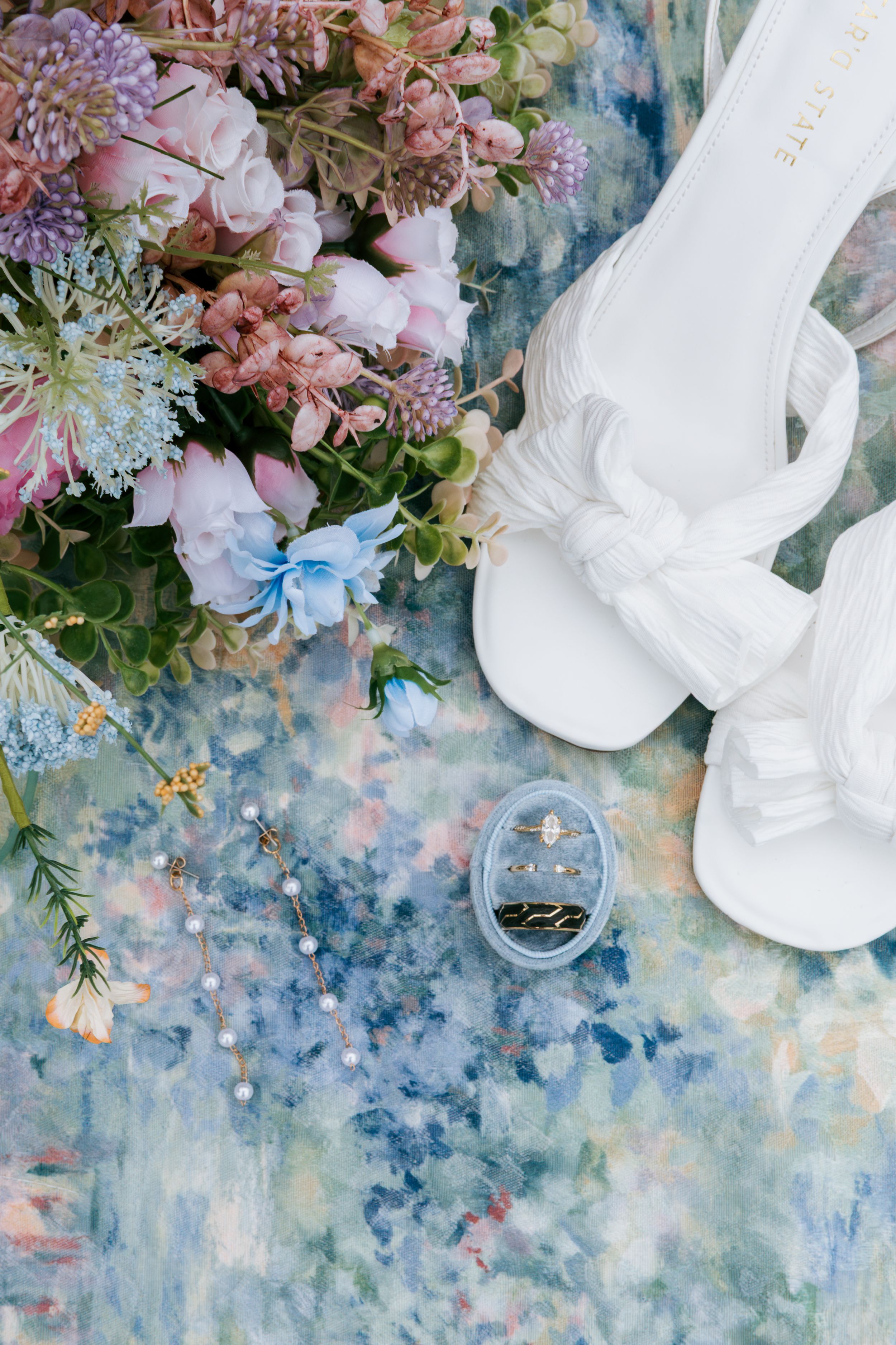 Bridal flat lay with white heels, floral bouquet, and wedding rings on artistic background, wedding photographer Asheboro NC