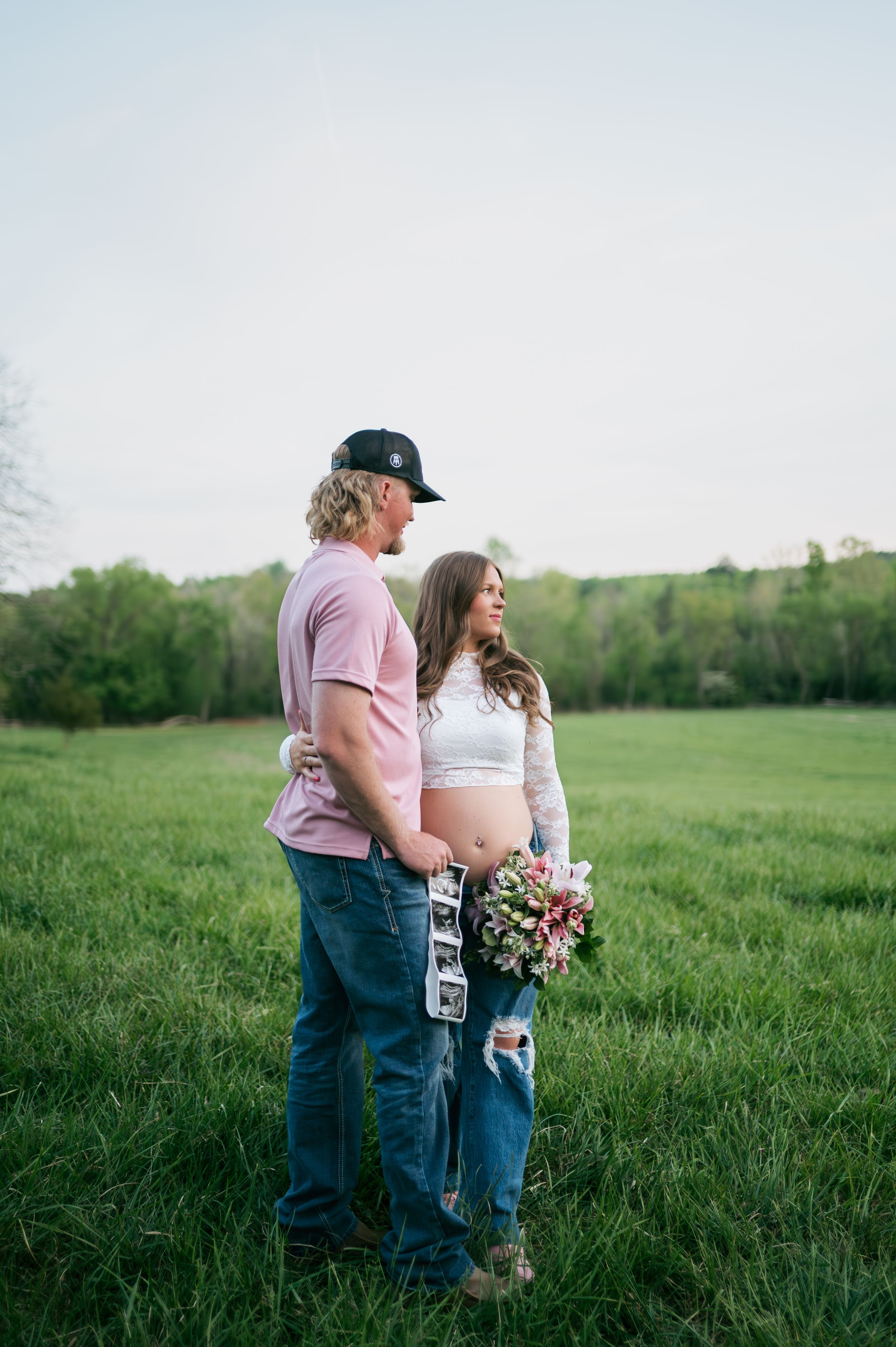 Couple posing in green field during maternity shoot, woman holding floral bouquet, maternity photographer North Carolina