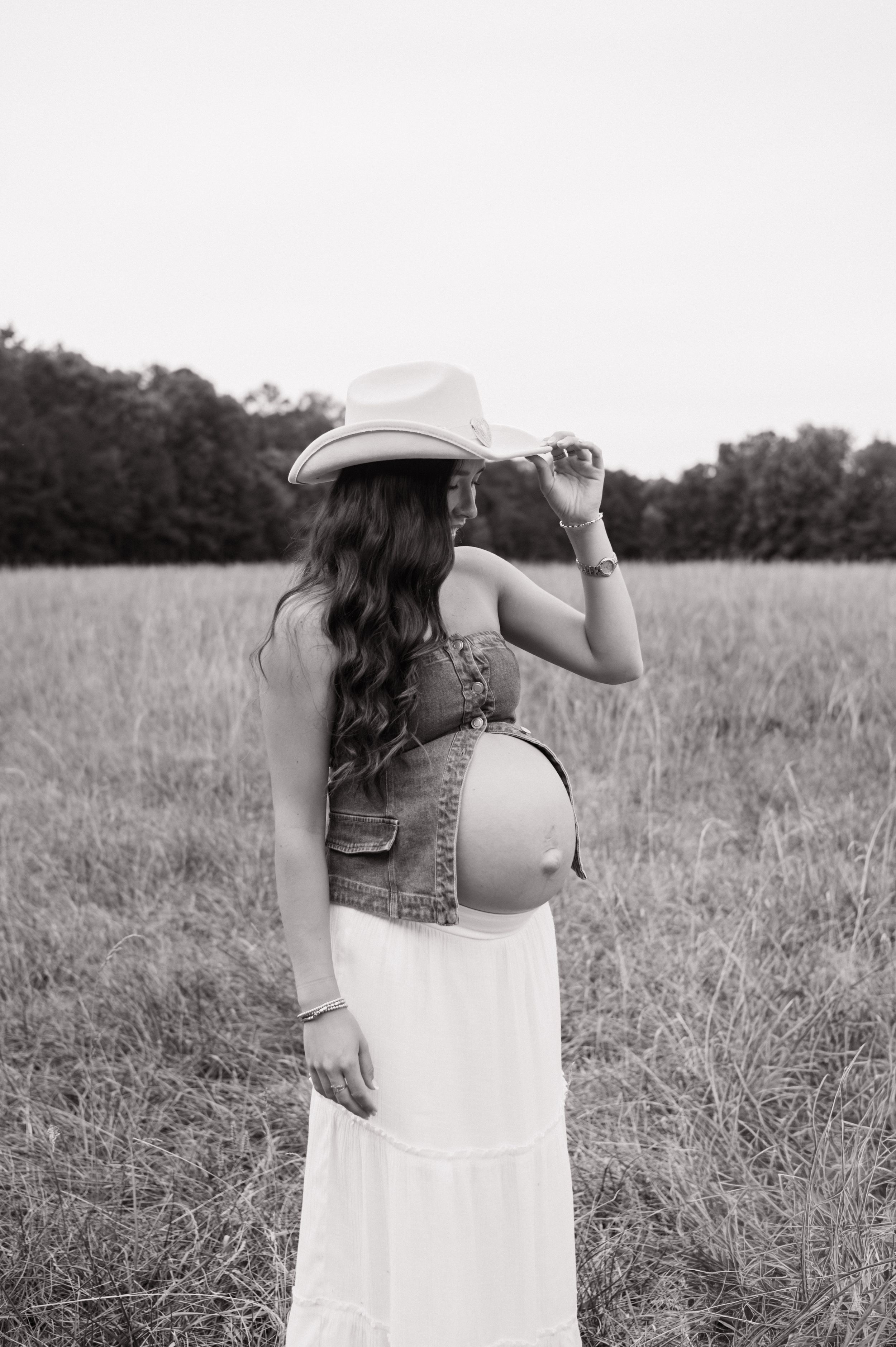 Black and white photo of pregnant woman in cowboy hat and white skirt in a field, maternity photographer North Carolina