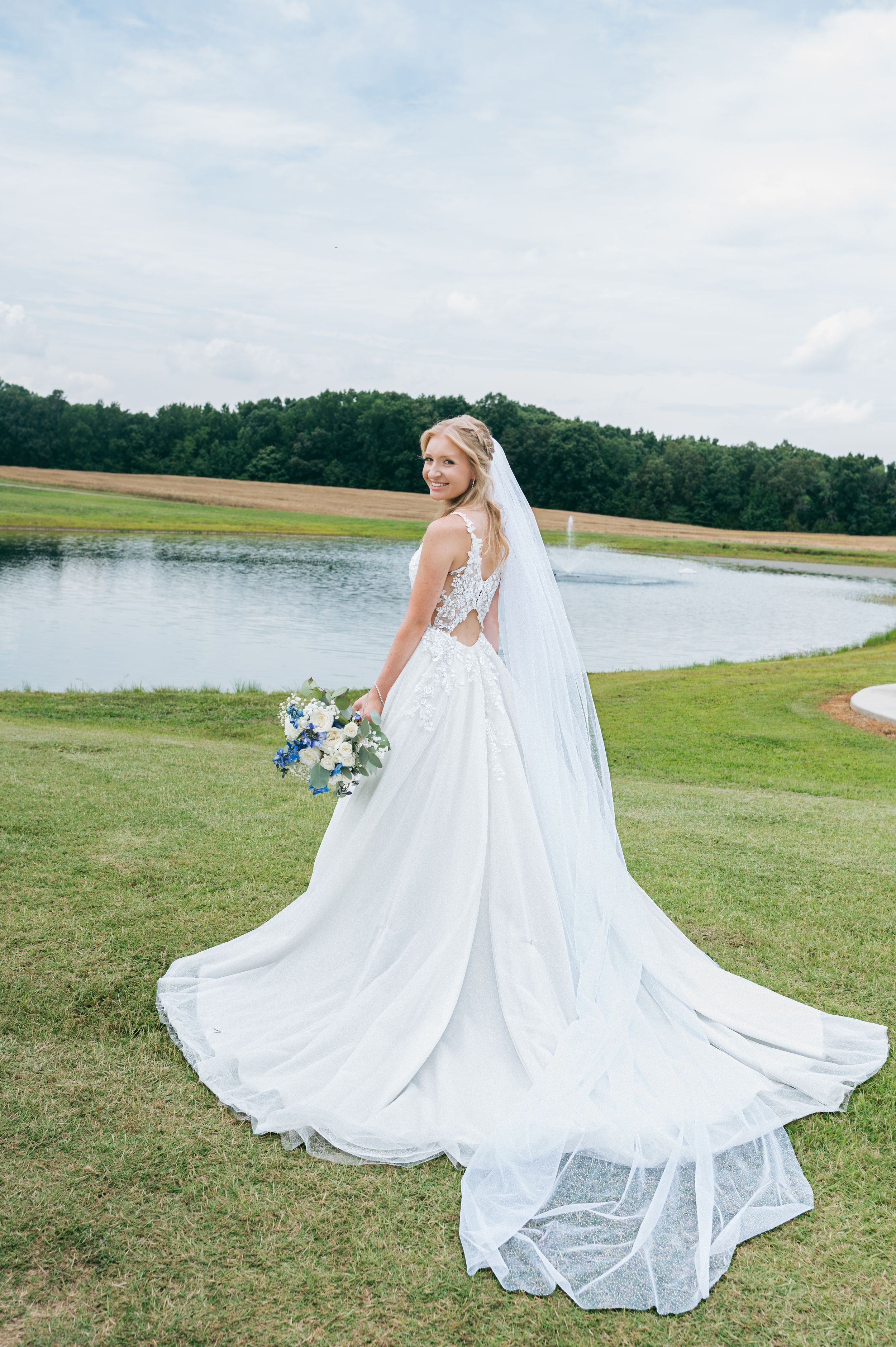 Bride in elegant gown with long veil holding bouquet beside a scenic pond, wedding photographer Greensboro NC