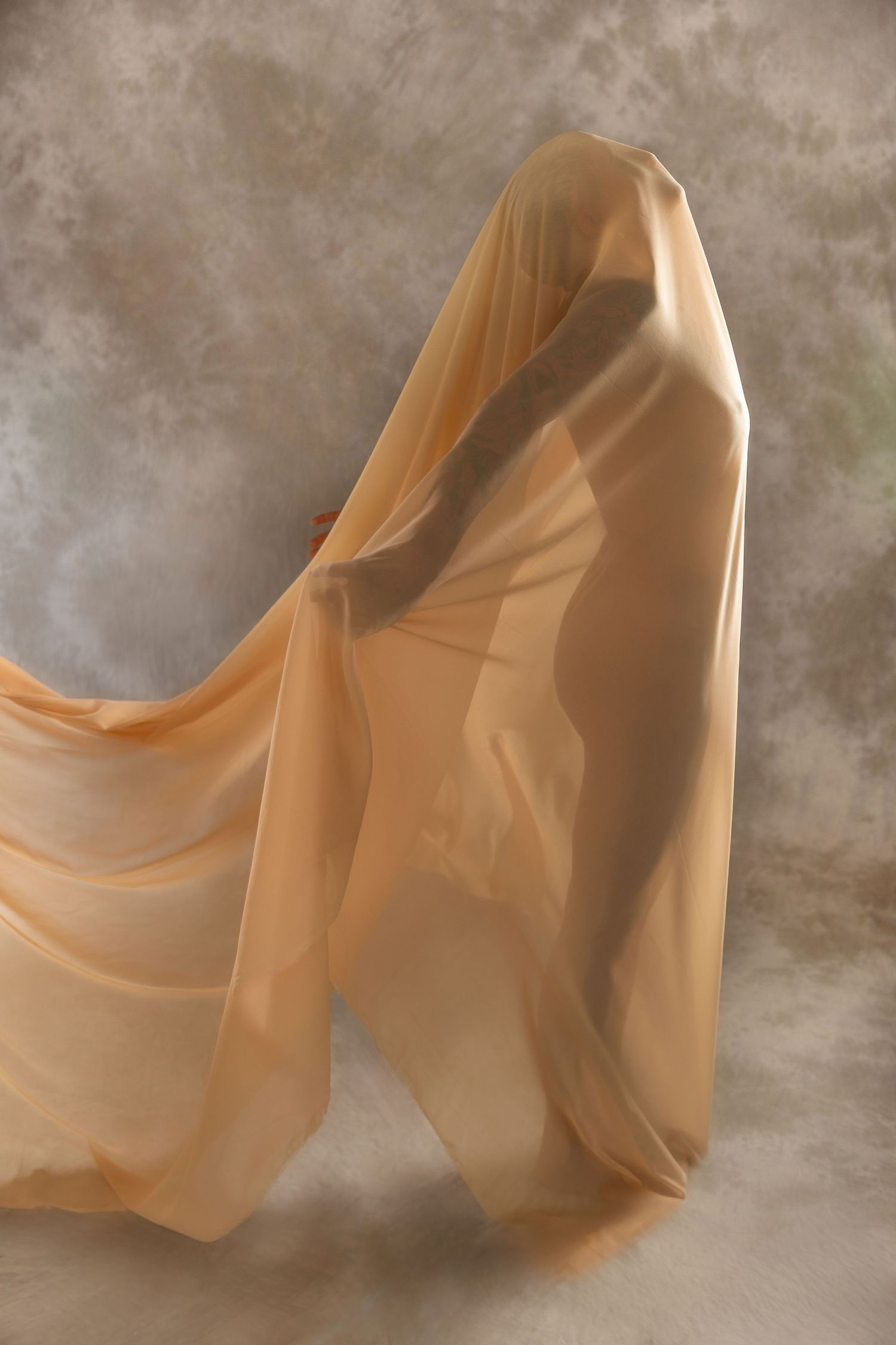 A figure draped in flowing sheer peach fabric against a muted studio background, creating an ethereal, ghostly silhouette.