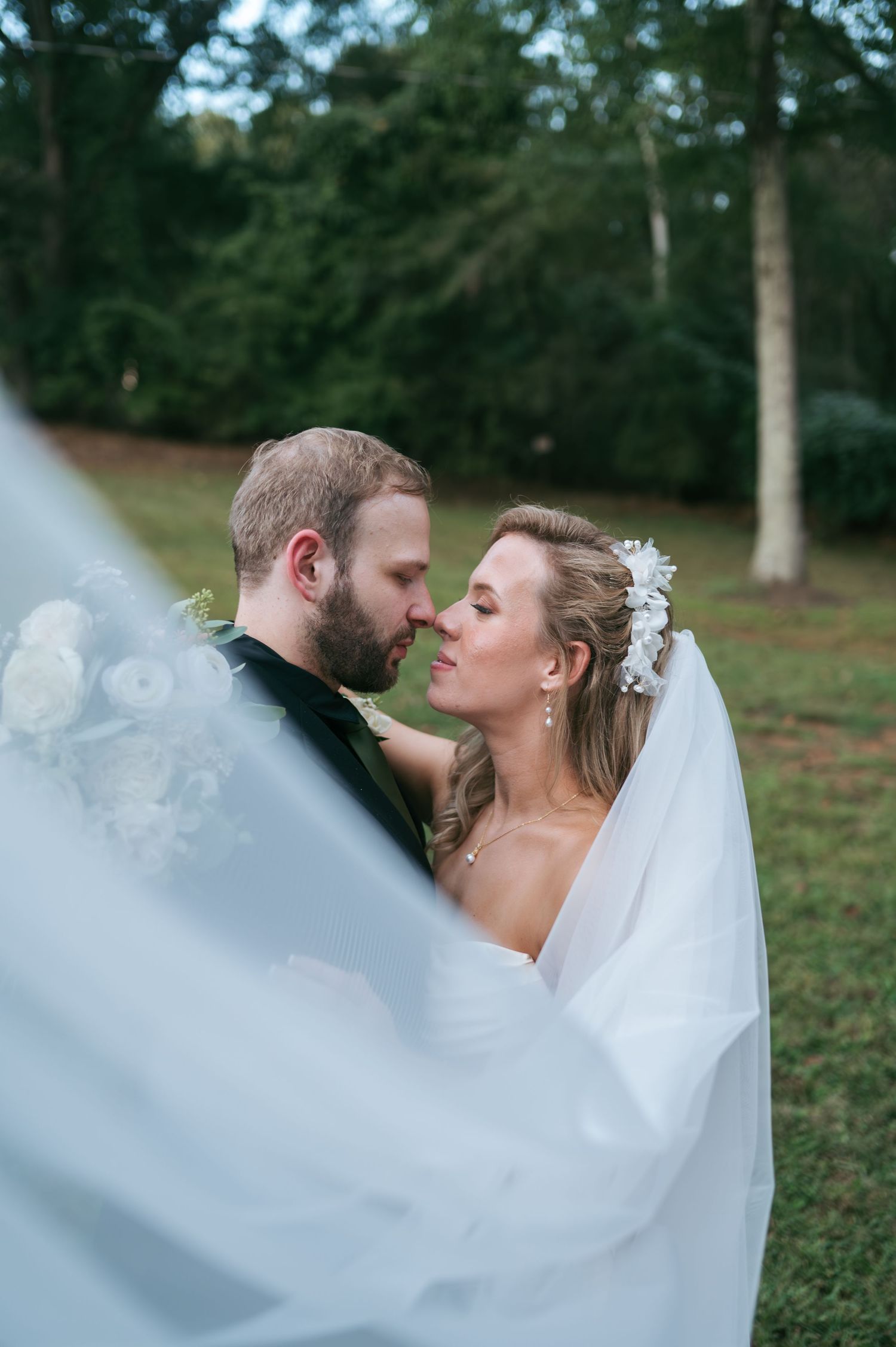 Bride and groom nearly kissing as her flowing veil wraps around them both in a lush green outdoor setting, wedding photographer Lexington NC