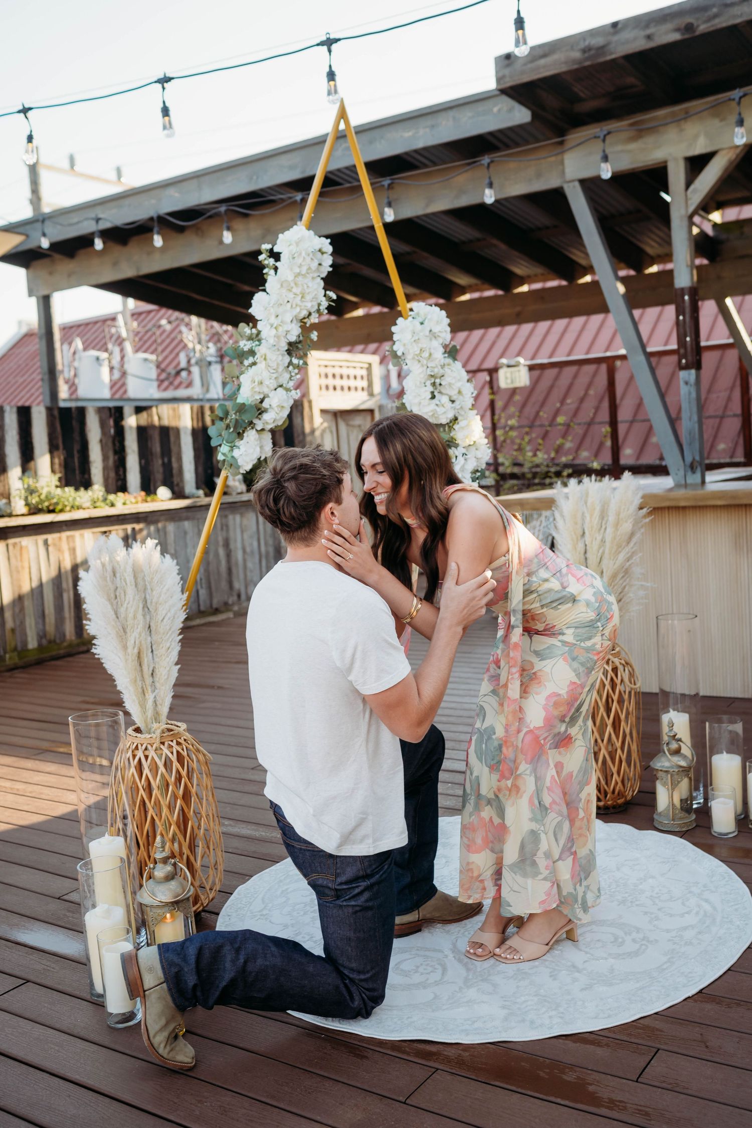 Man proposing on one knee to woman on rooftop deck with floral arch, candles, and pampas grass decor.