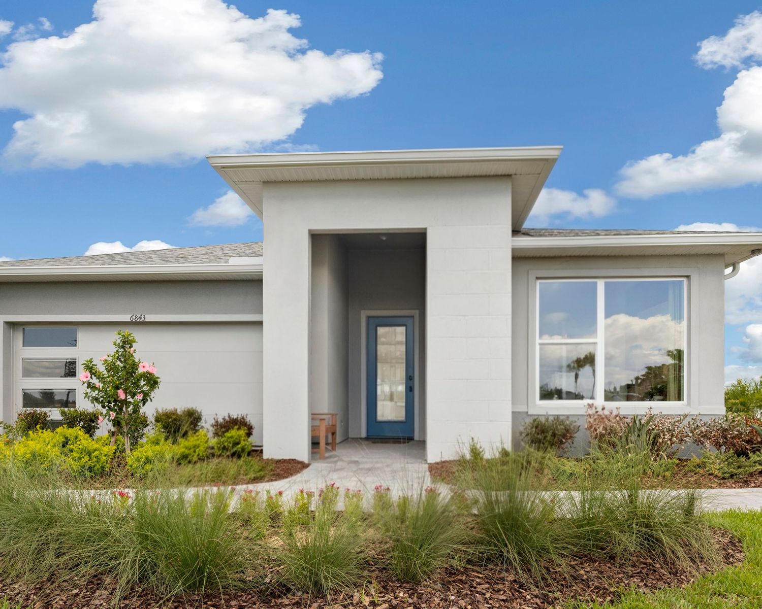 Modern single-story home with gray stucco exterior, blue front door, large windows, and lush green landscaping under a blue sky.