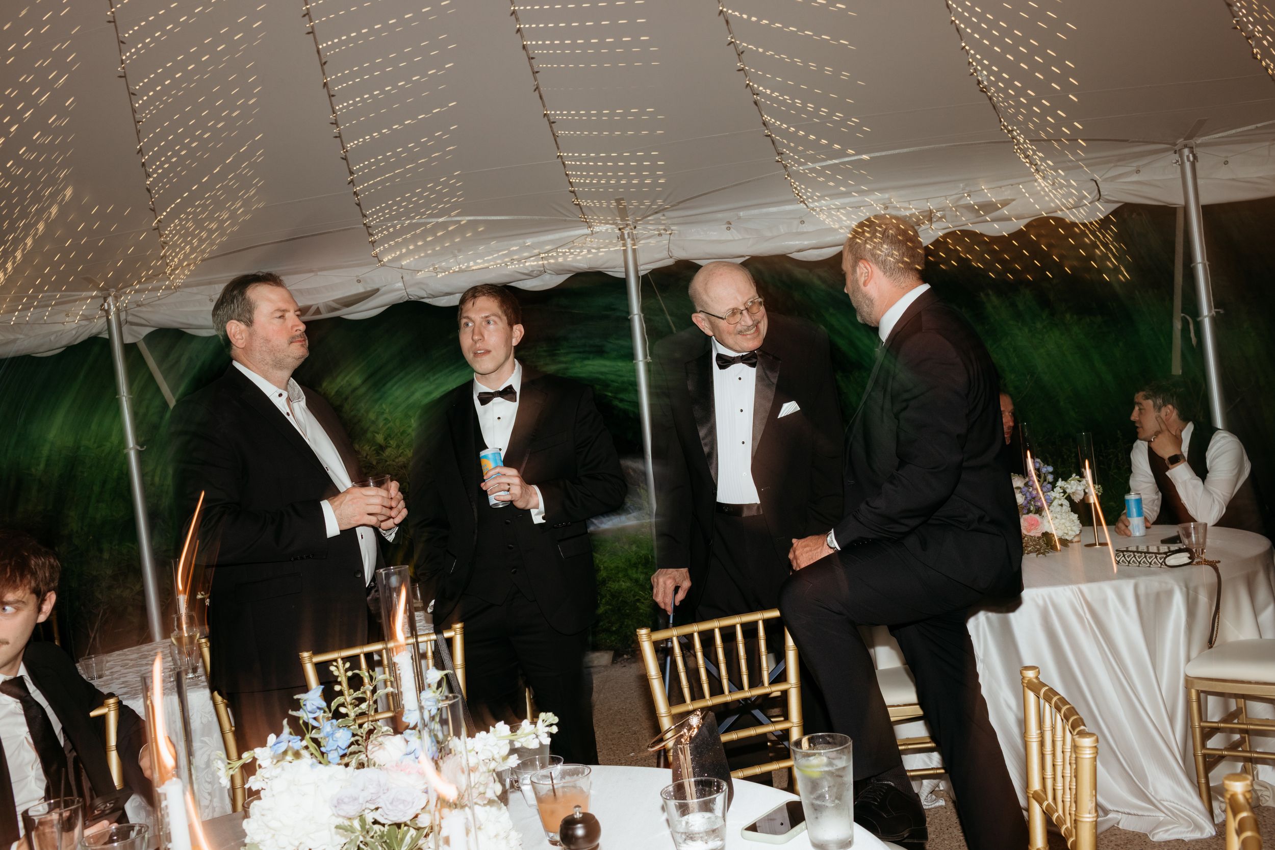 Four men in tuxedos socializing under a elegant white tent at a formal wedding reception.