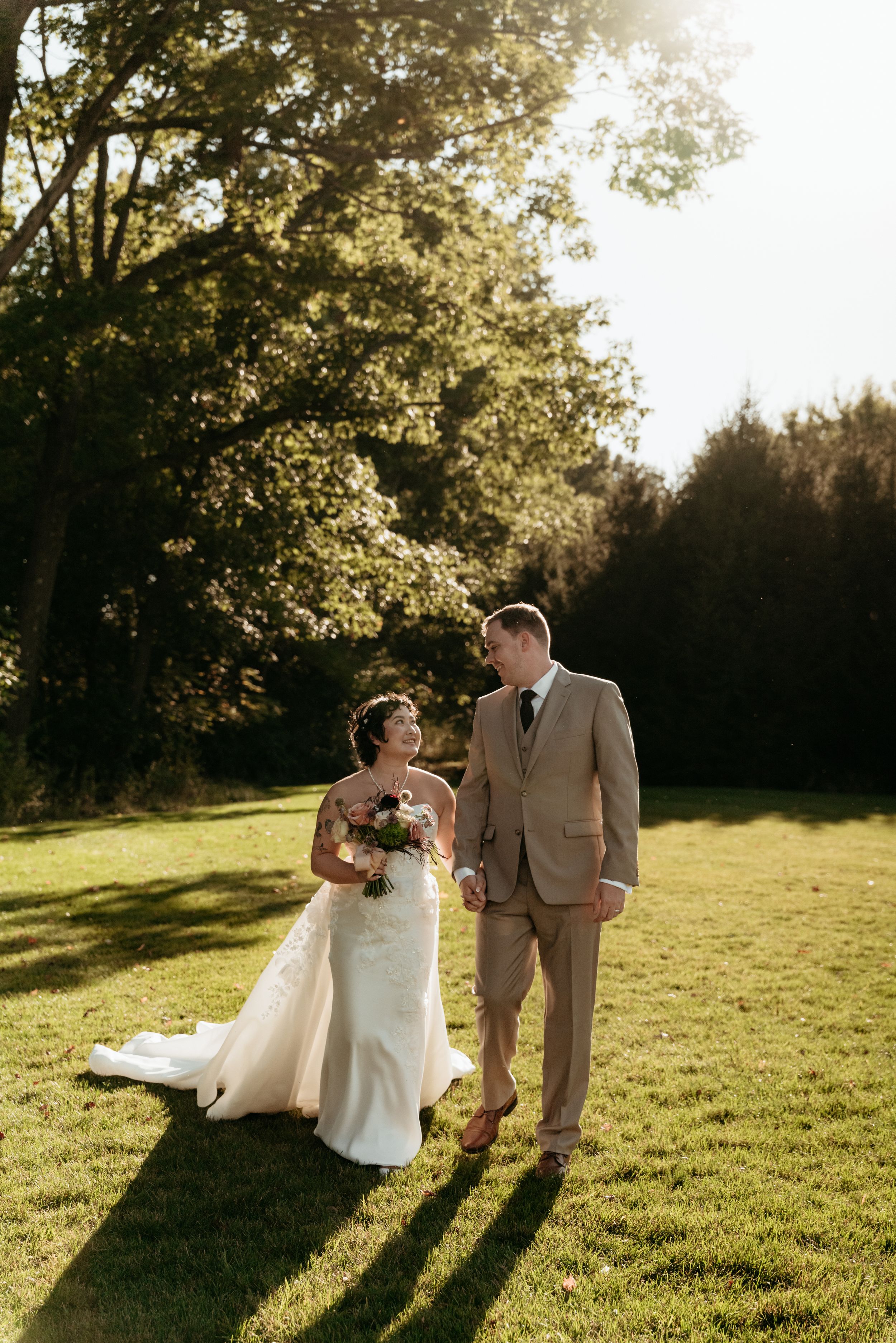Bride and groom walking hand in hand on lush green lawn, bride in white gown holding bouquet, groom in tan suit, golden hour.