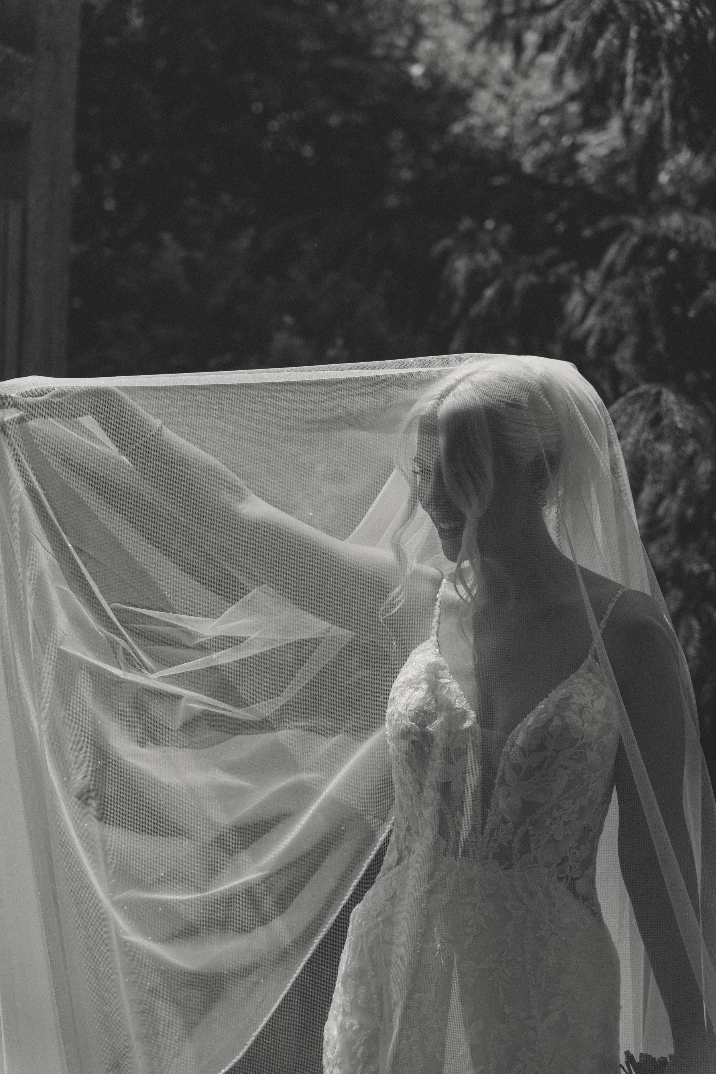 Black and white photo of a bride in a lace gown lifting her flowing veil outdoors with trees in the background.