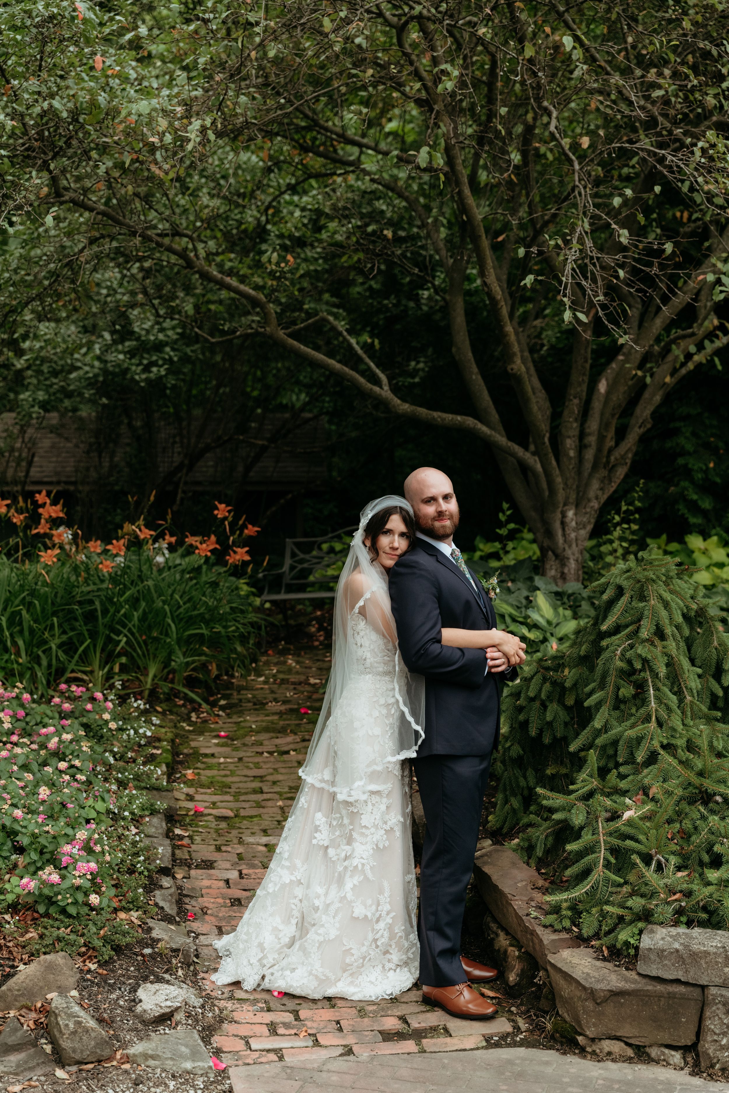 Bride in lace gown and veil stands with groom in navy suit on a brick path surrounded by lush garden greenery.