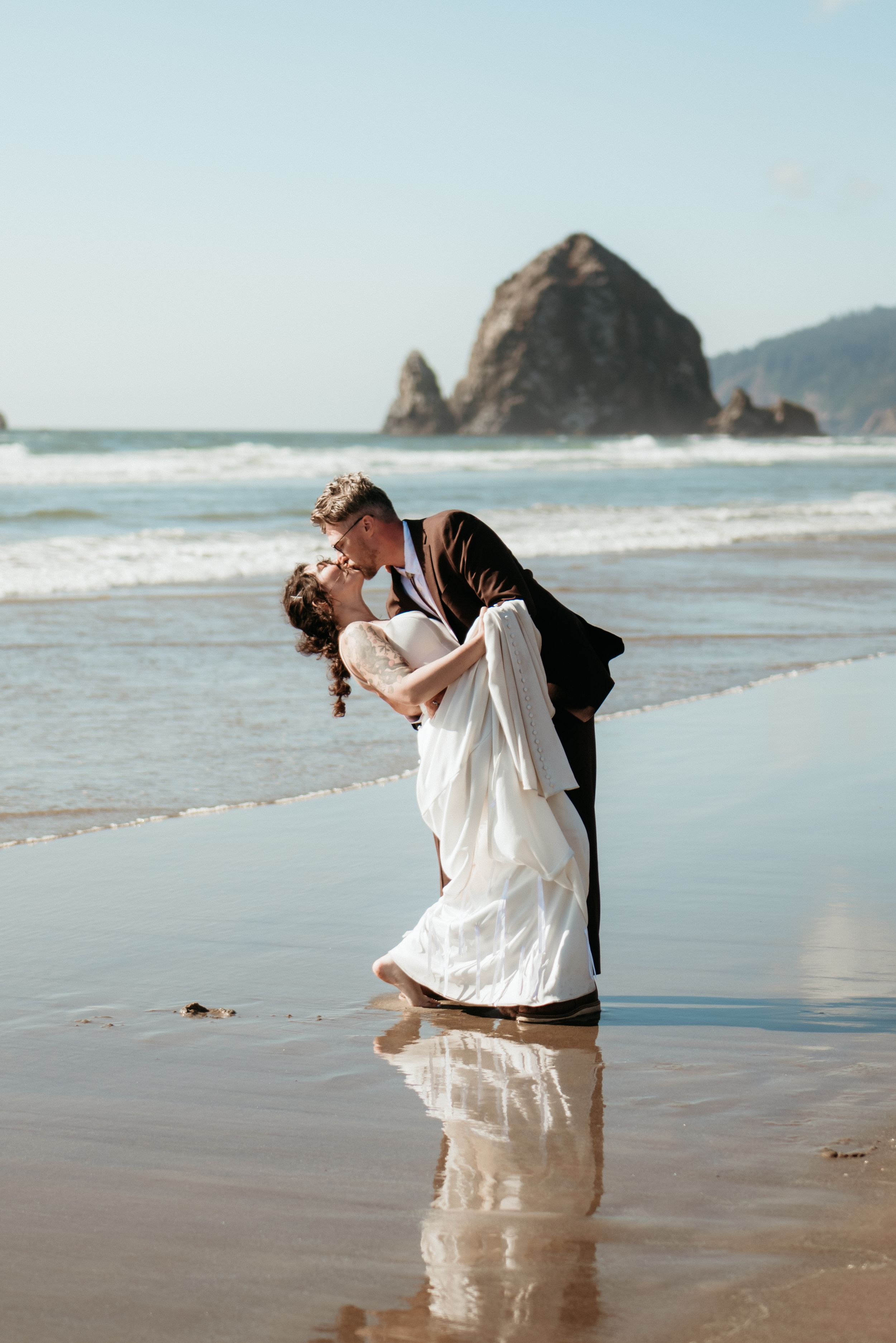 Bride and groom share a romantic dip kiss on a wet beach with Haystack Rock in the background.