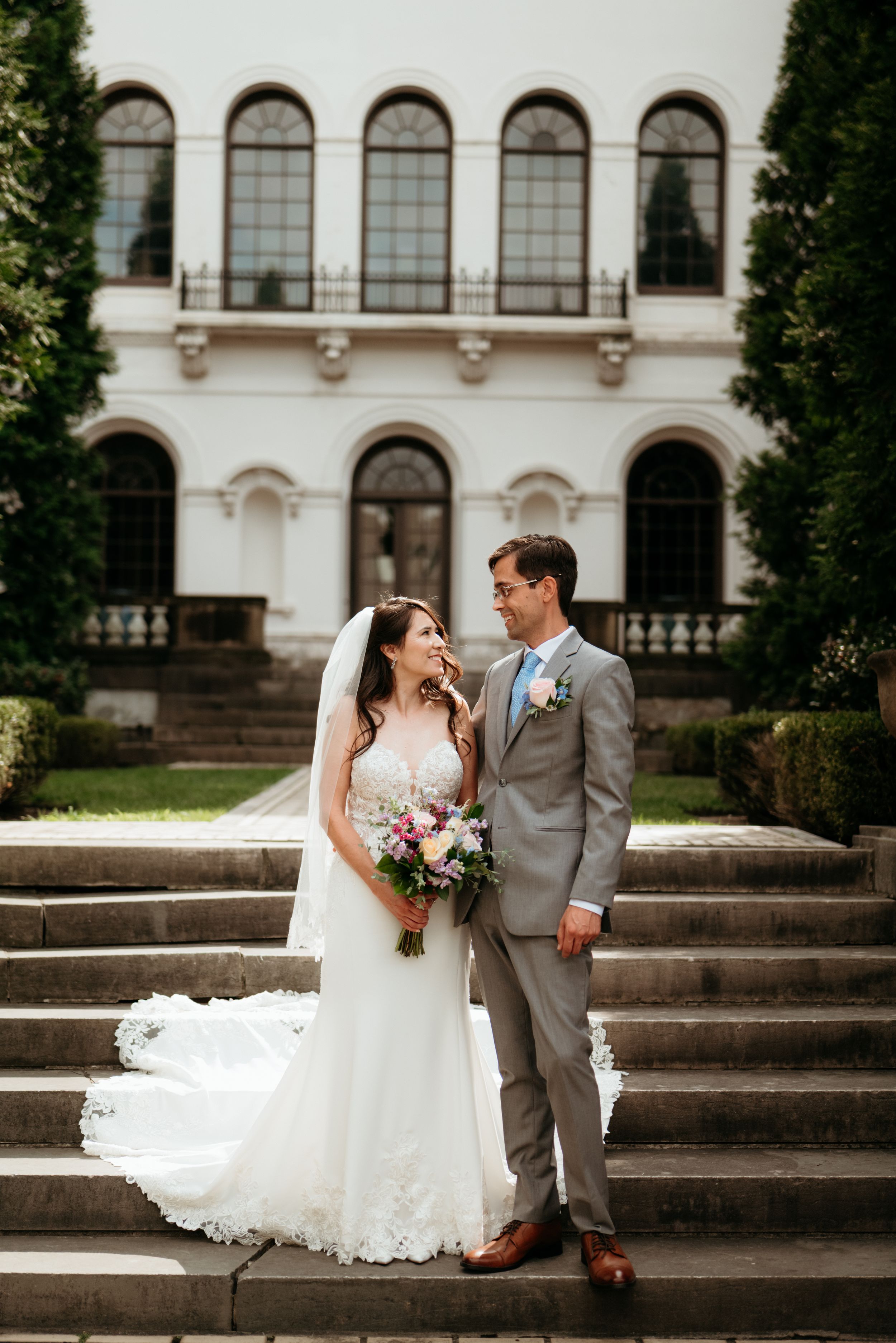 Bride and groom stand on stone steps before elegant white mansion, bride holding colorful bouquet in lace wedding gown.