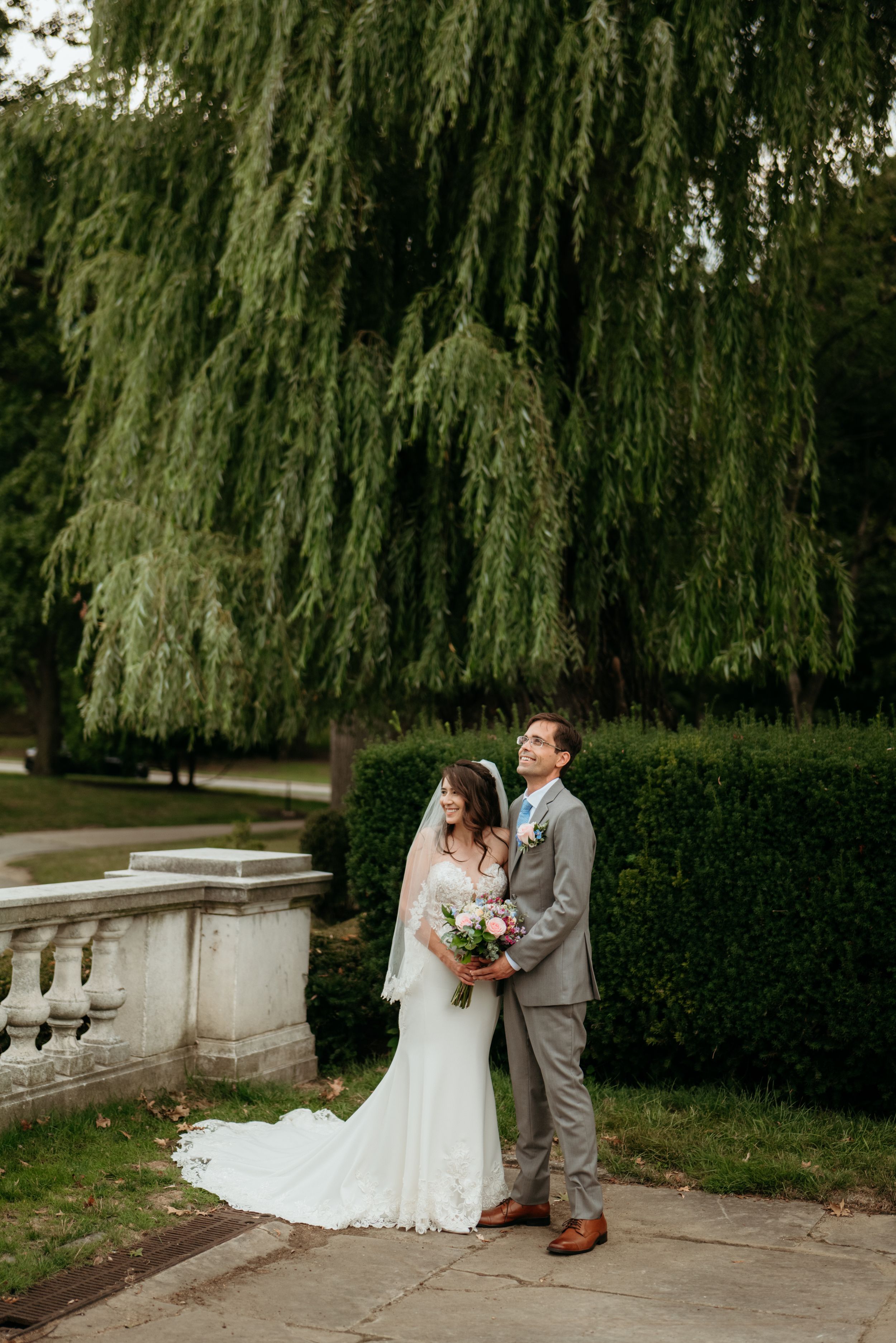 Bride and groom pose together under a weeping willow tree in a lush garden, bride holding a colorful bouquet.