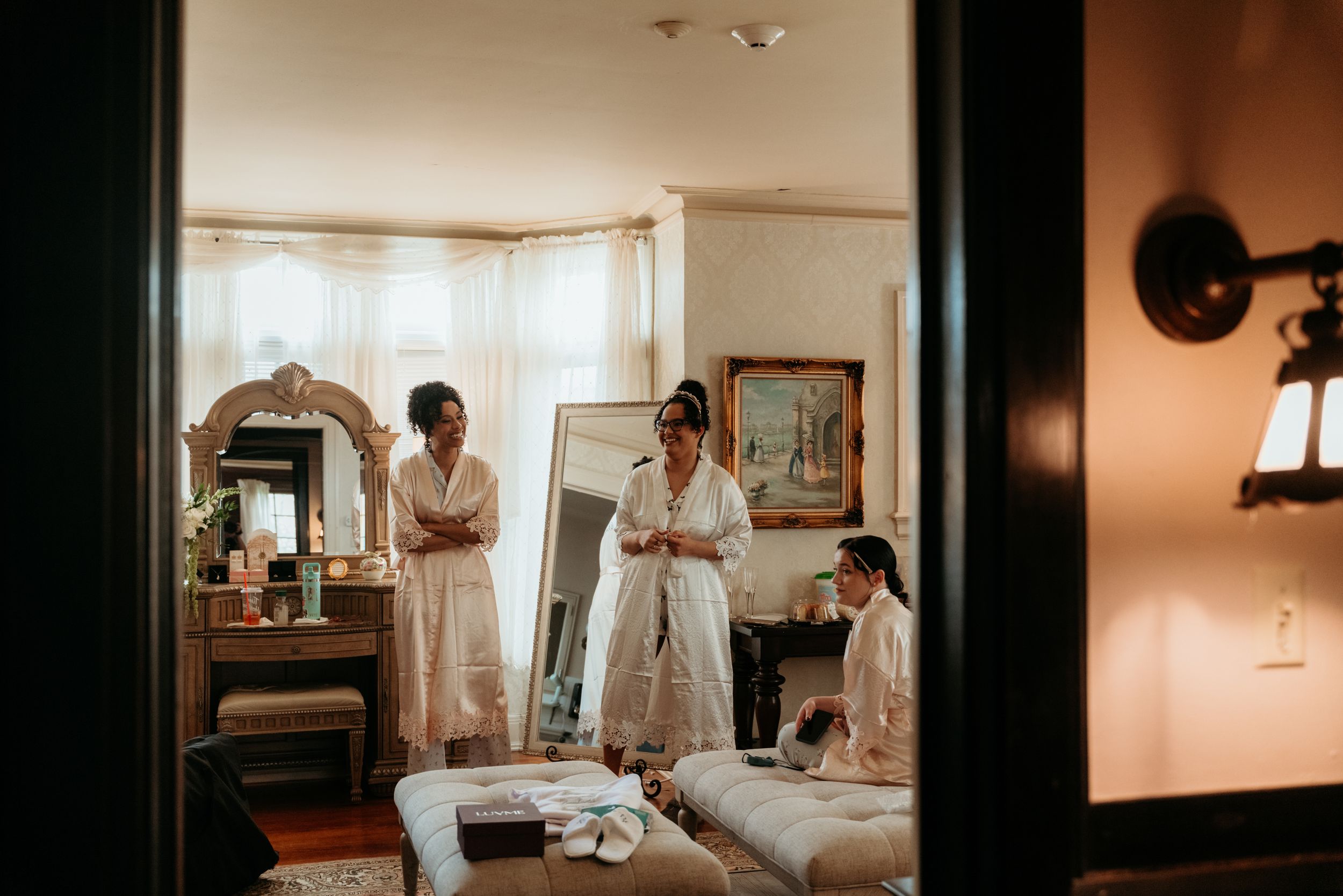 Bridesmaids in white robes preparing in an elegant vintage bedroom with a mirror and vanity before a wedding.