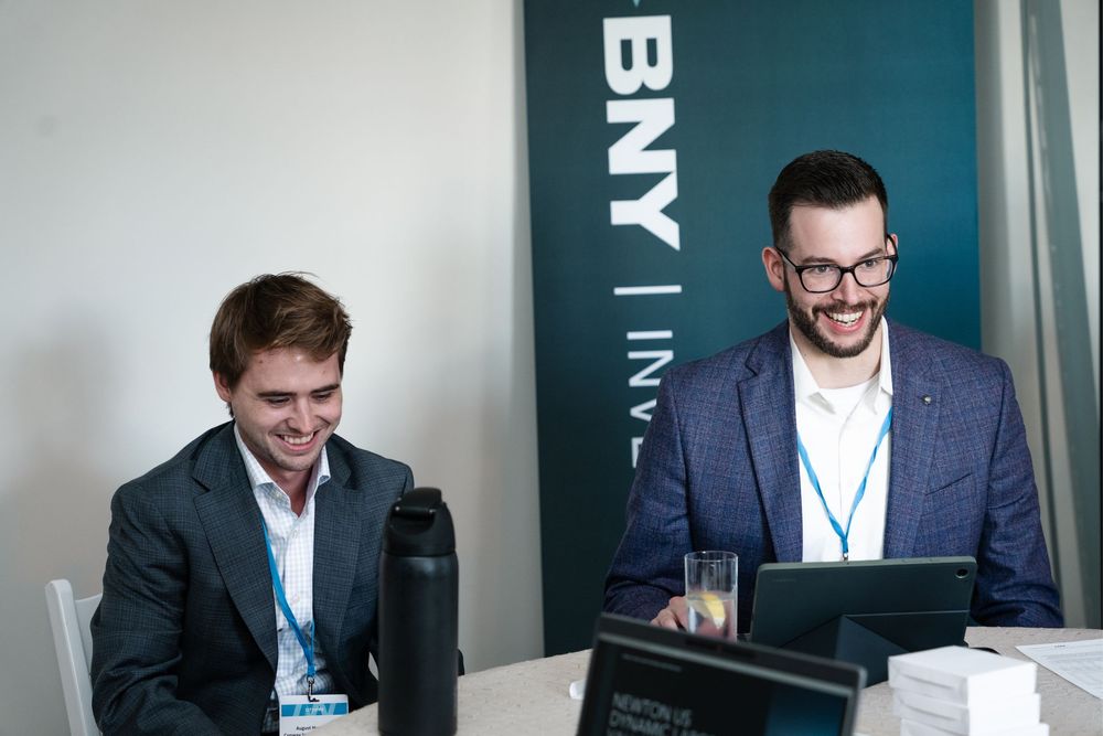 Two smiling businessmen in suits wearing lanyards sit at a desk in front of a BNY Investments banner.