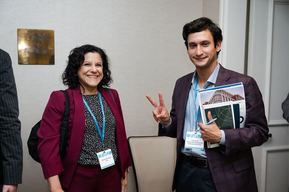 Two conference attendees smiling and posing for photo, woman in burgundy blazer with lanyard.