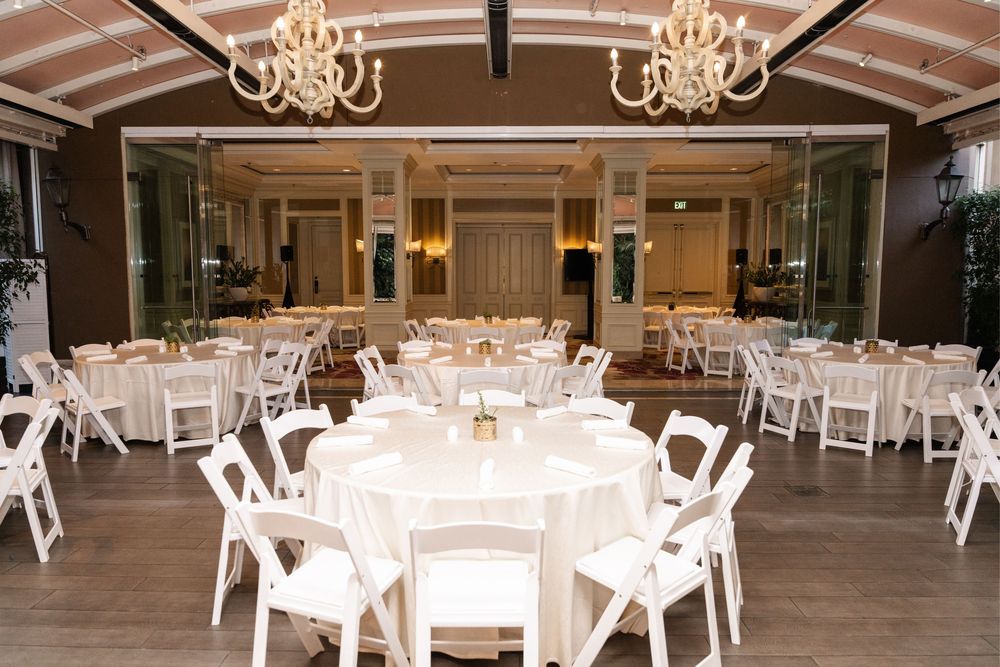 Elegant wedding reception hall with round tables, white chairs, and chandeliers on wood ceiling.
