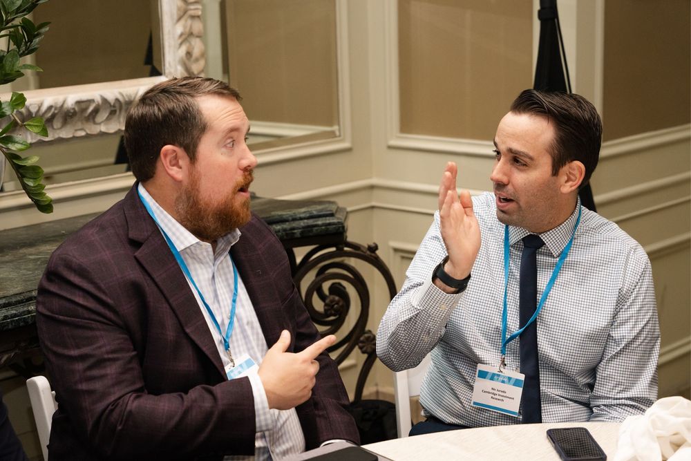 Two business professionals in suits engage in animated conversation at a conference table wearing name badges.