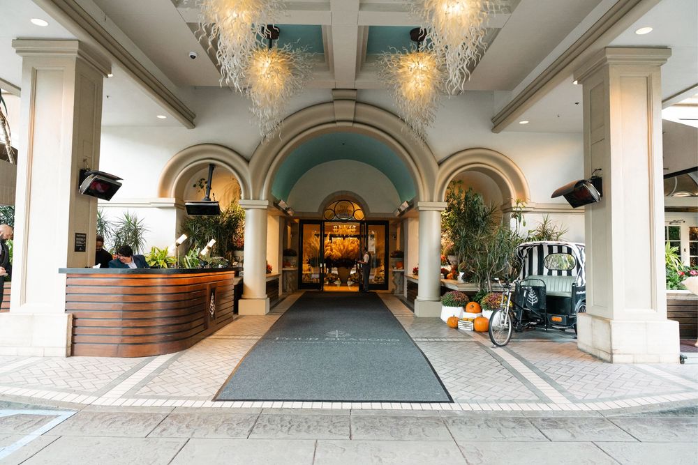 Elegant hotel lobby entrance with arched doorways, ornate chandeliers, marble columns, and lush greenery on a tiled walkway.