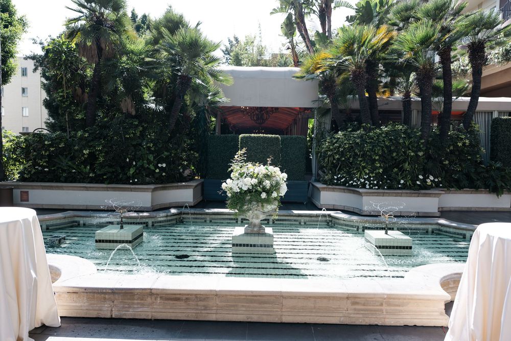 Elegant outdoor fountain courtyard with white floral centerpiece, surrounded by lush palm trees and manicured hedges.