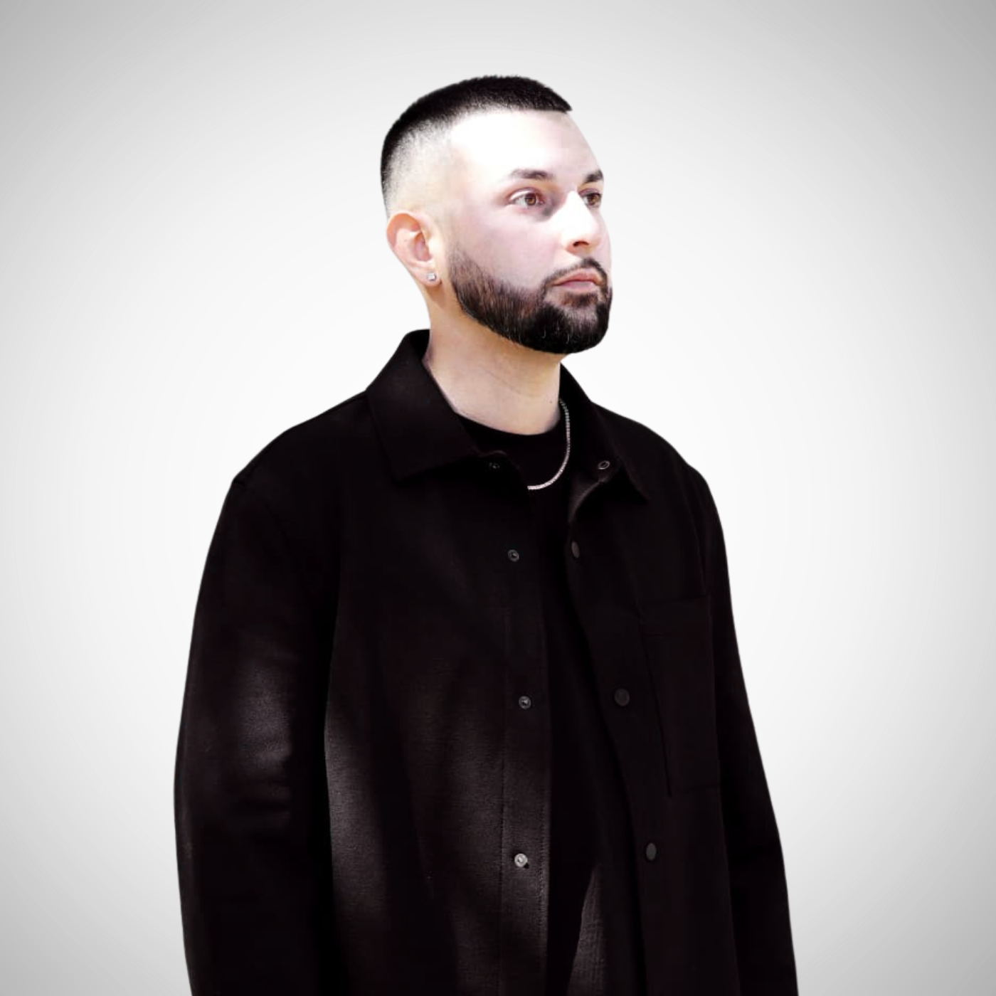 A man with a beard and short hair wearing a black oversized jacket and chain necklace poses against a gray background.