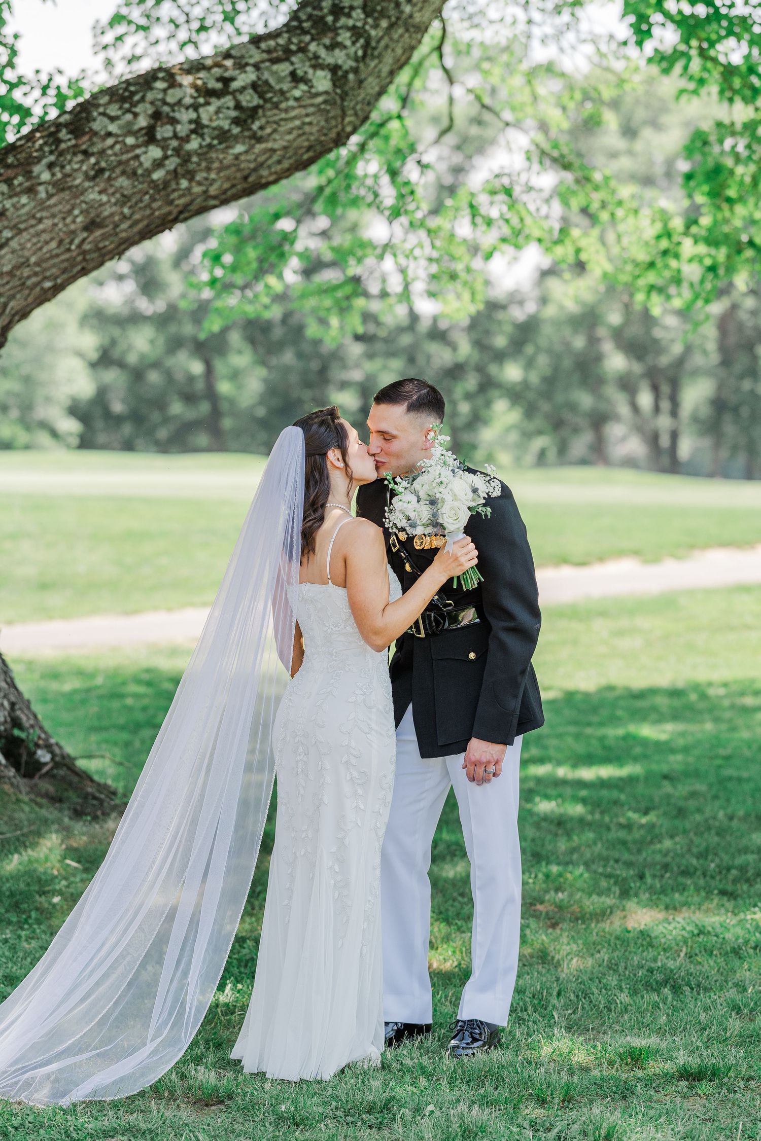 A bride and groom share a kiss under a large tree on lush green grass, she in a white gown with veil, he in military dress.