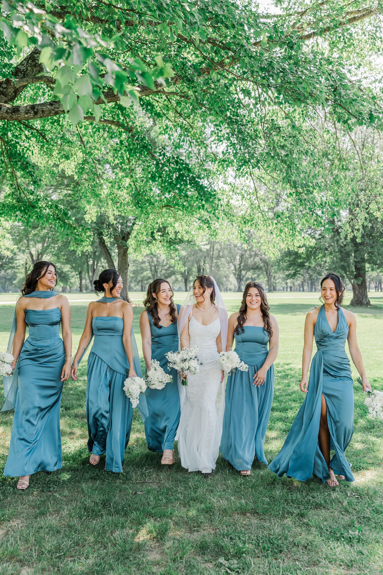 Bride in white gown walks with five bridesmaids in dusty blue dresses through a lush green park.