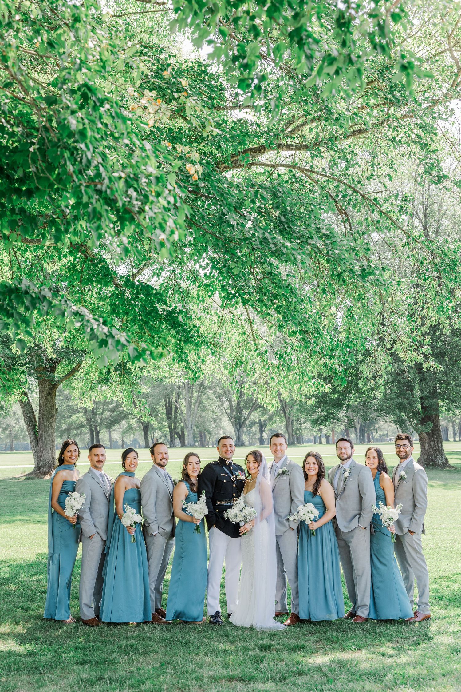 Wedding party in dusty blue gowns and gray suits posing outdoors under lush green trees with bride and groom centered.