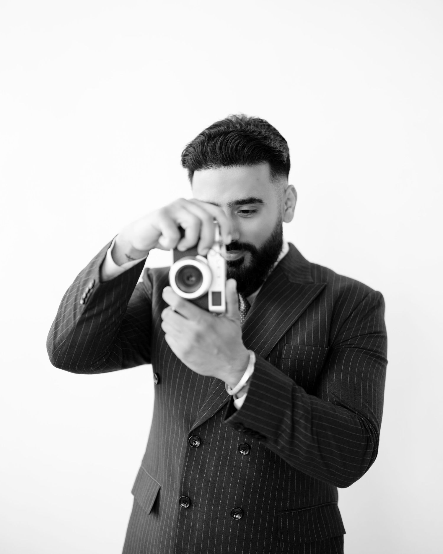 Black and white photo of a bearded man in a pinstripe suit holding a vintage camera up to his eye.