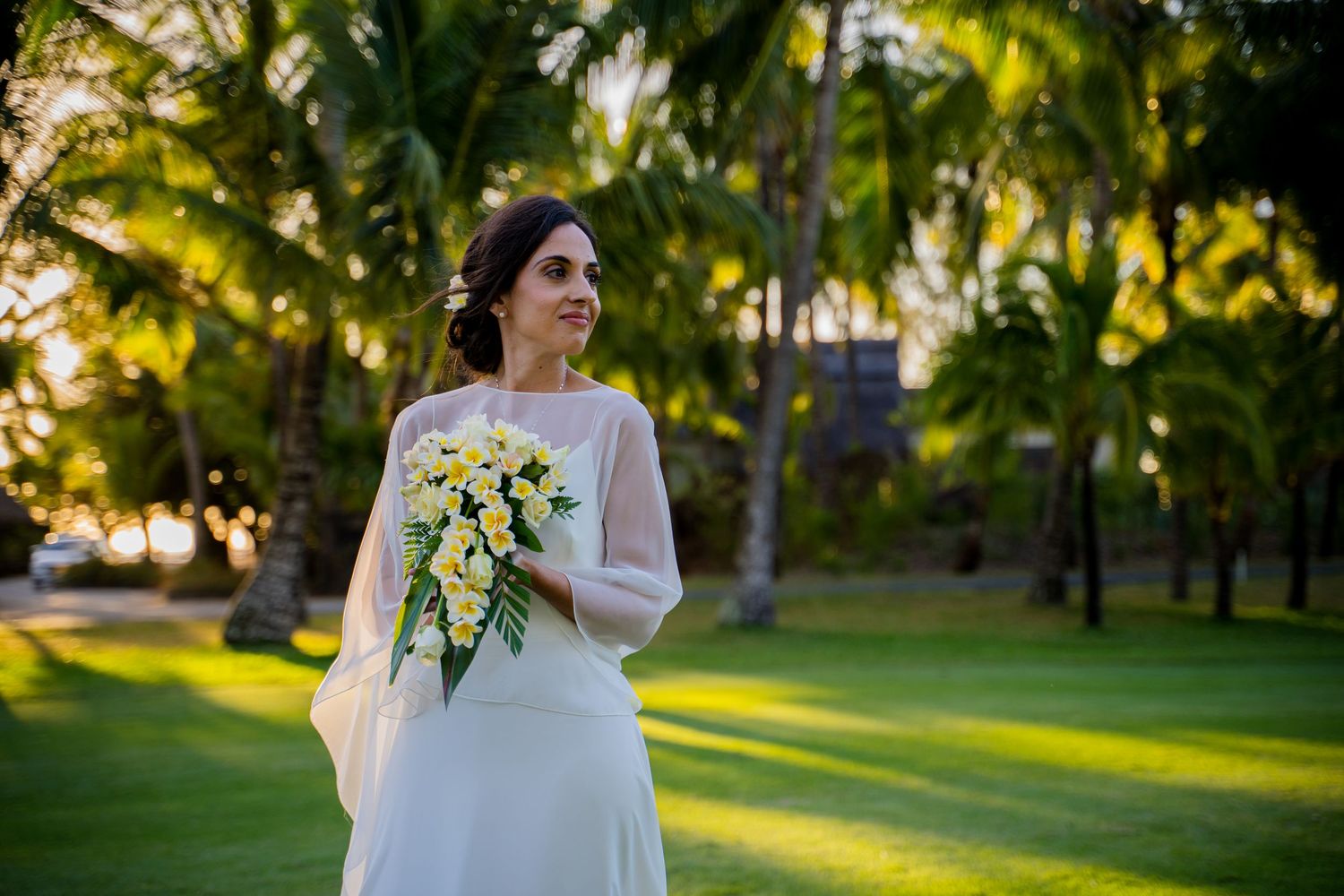 Bride in white gown holding yellow floral bouquet standing on lush green lawn surrounded by tropical palm trees at sunset.
