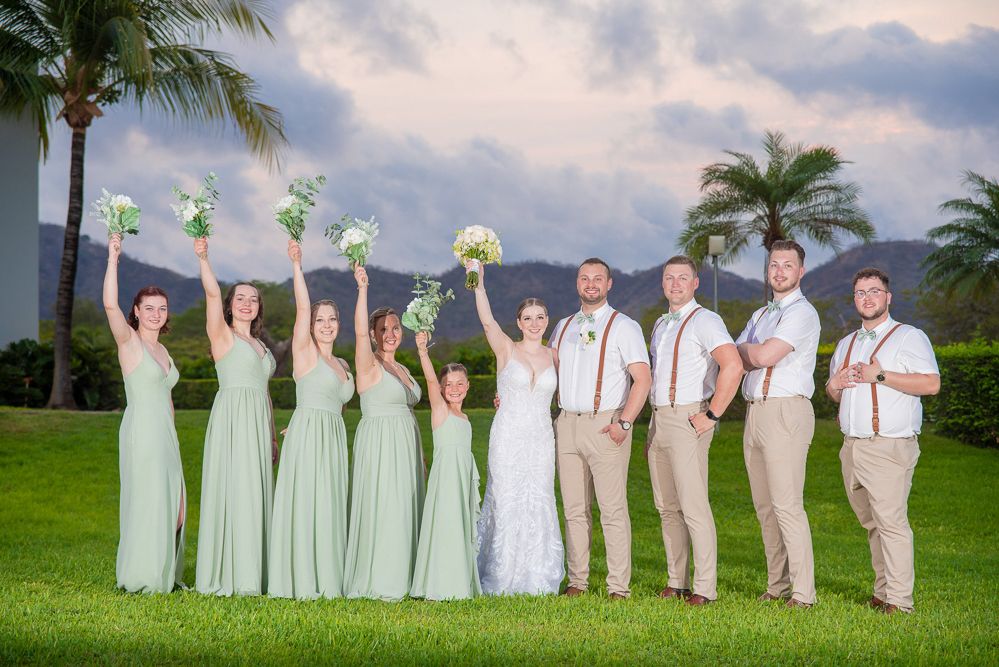 Wedding party poses outdoors with bridesmaids in sage green gowns and groomsmen in khaki, raising bouquets against a tropical backdrop.